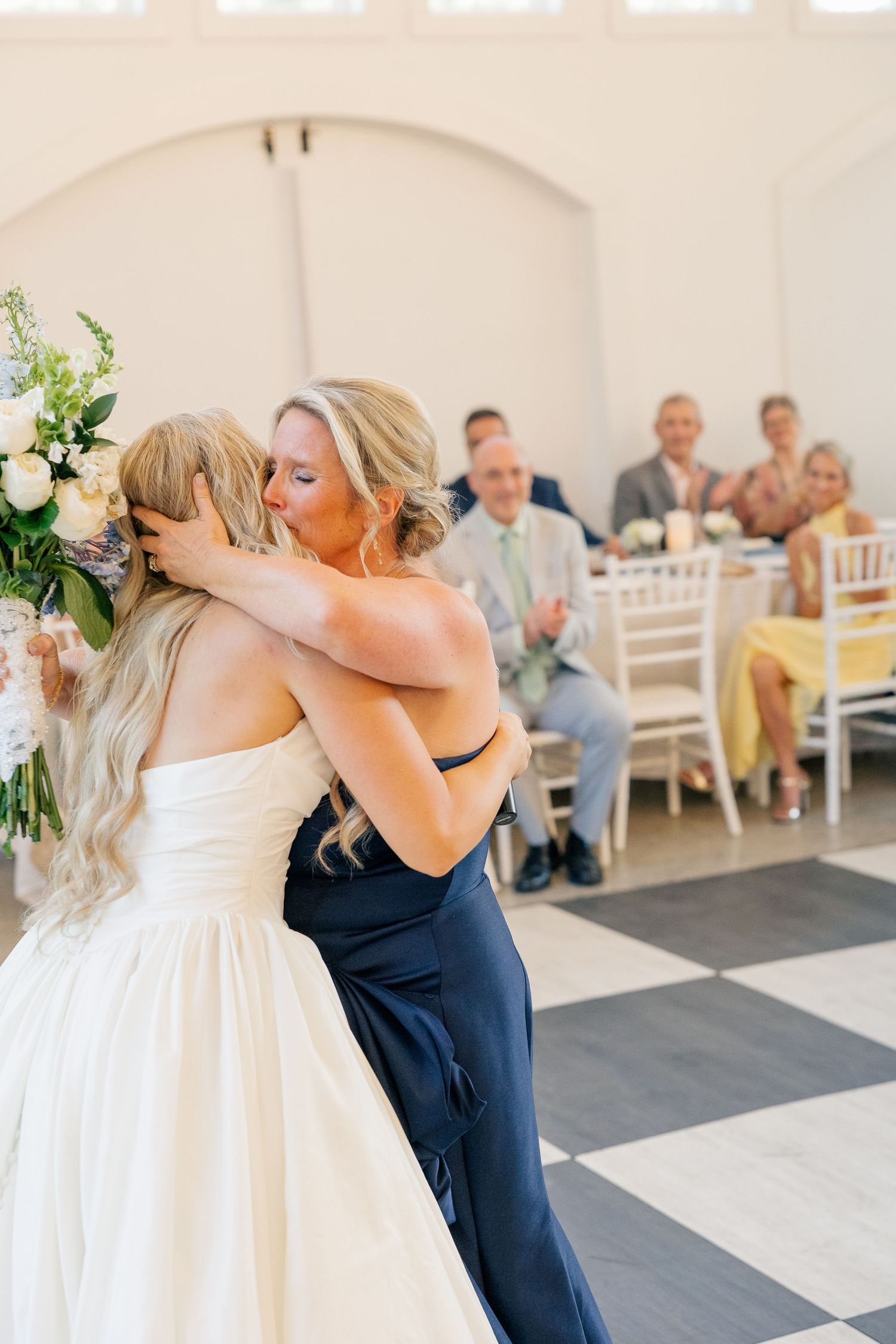 Bride in strapless white gown shares heartfelt hug with bridesmaid in navy dress on dance floor.