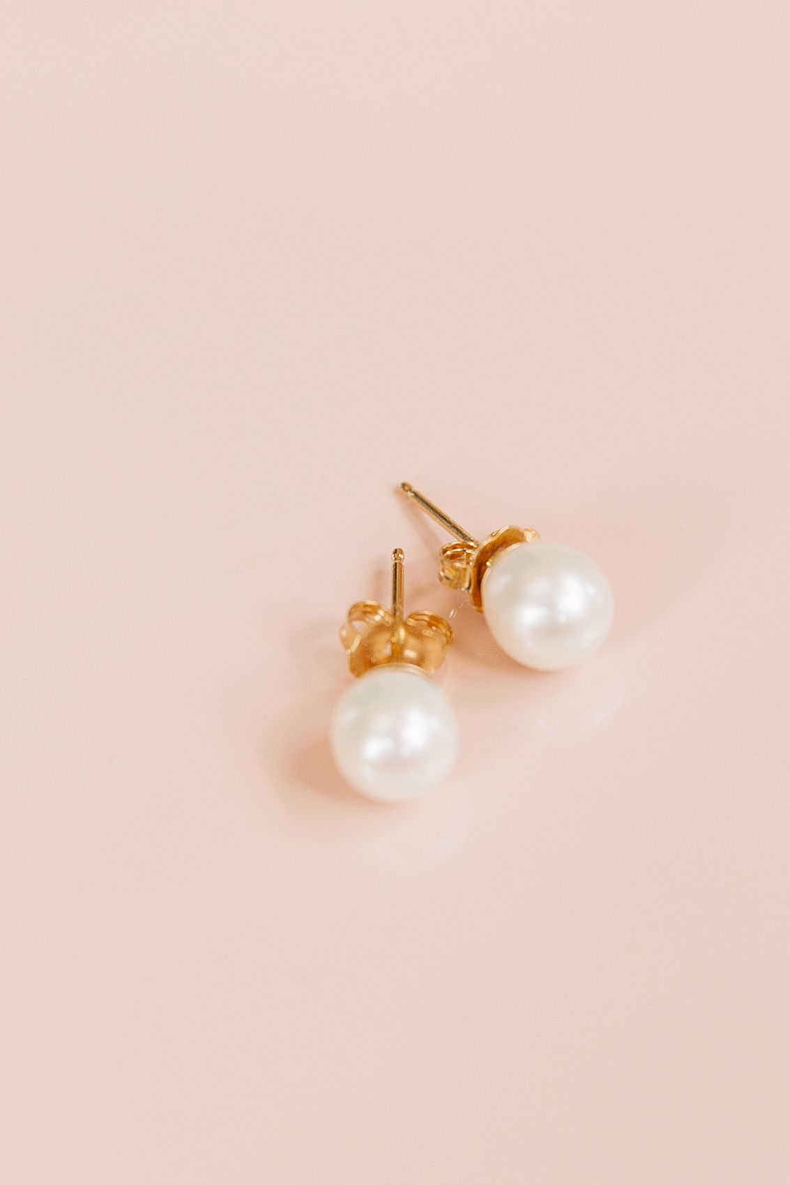 Gold pearl stud earrings with white freshwater pearls on a soft pink background.
