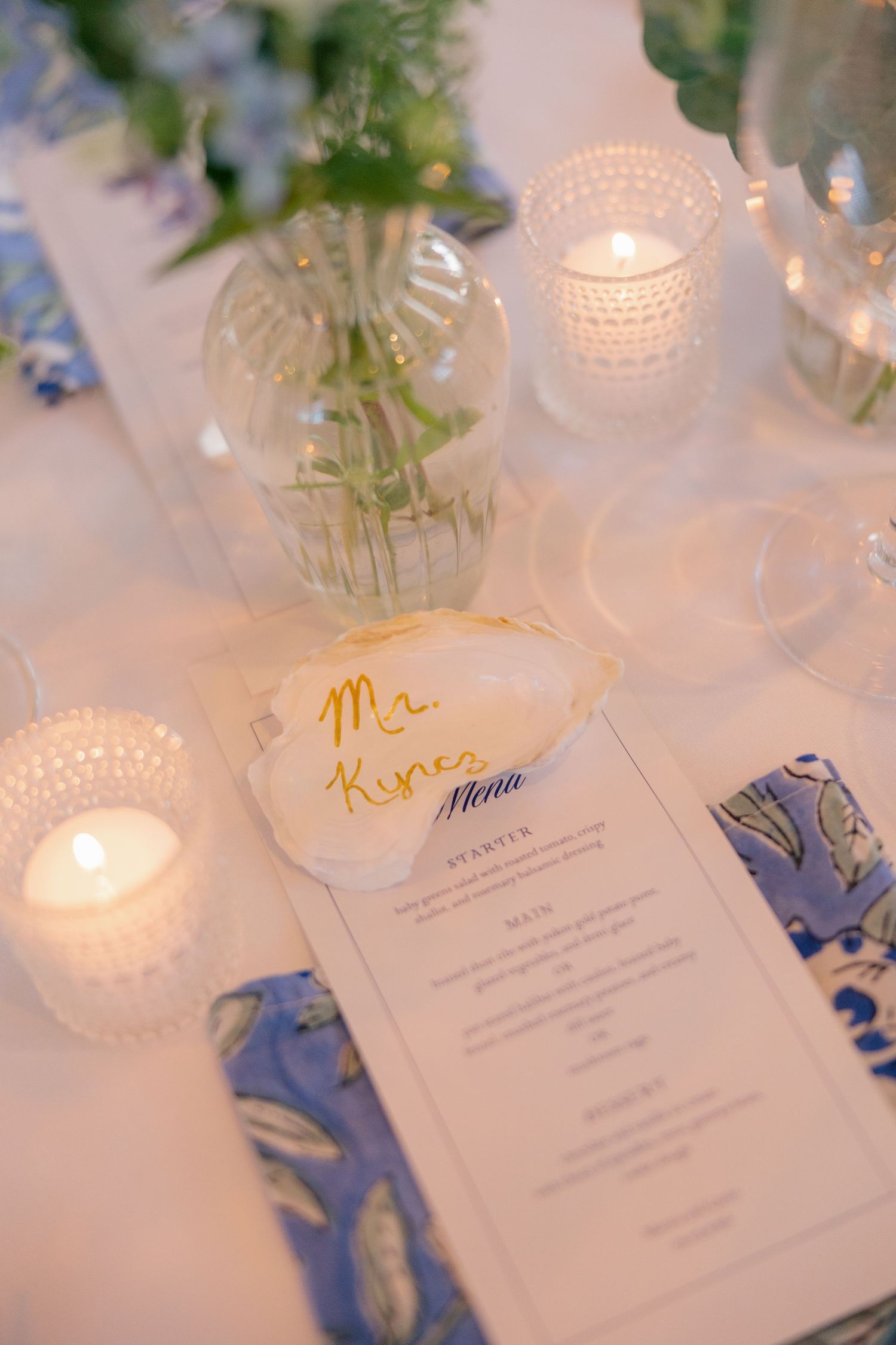 Elegant wedding place setting with shell name card, menu, blue napkin, and glowing candles on white linen.