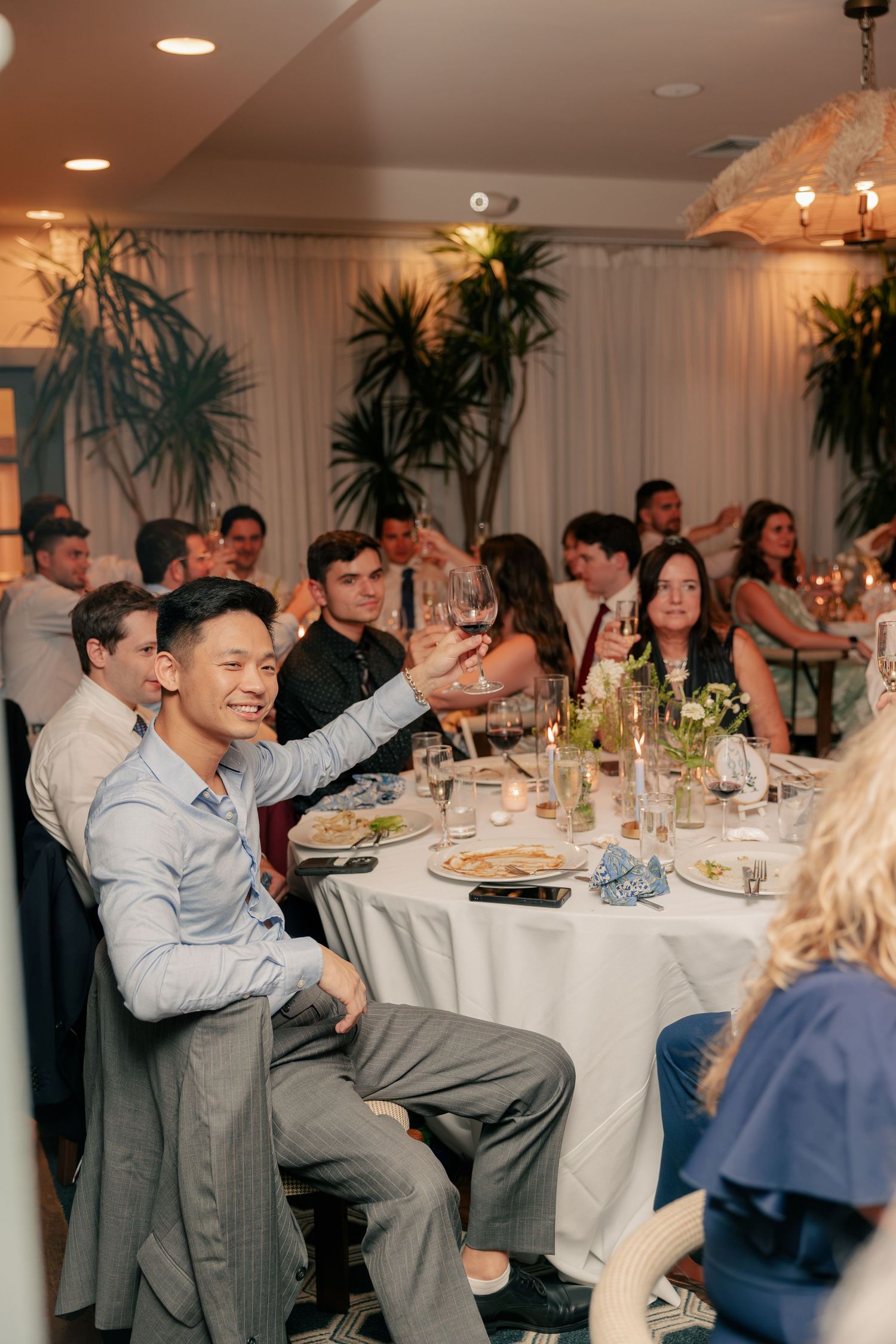 Wedding guests raising glasses in a toast at an elegant indoor reception dinner with white tablecloths.