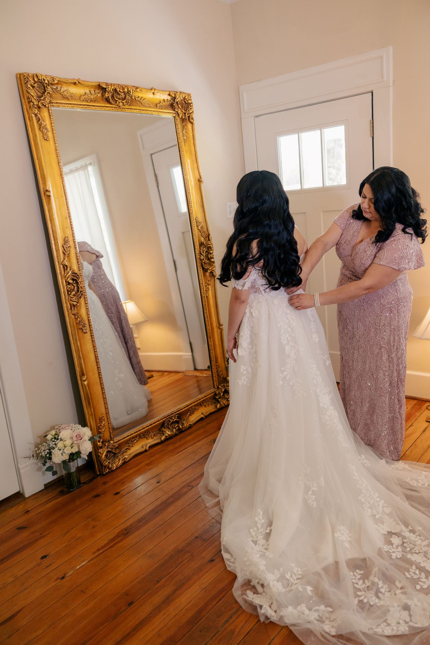 Elegant wedding dress with lace details and long train being prepared in dressing room with gold mirror.
