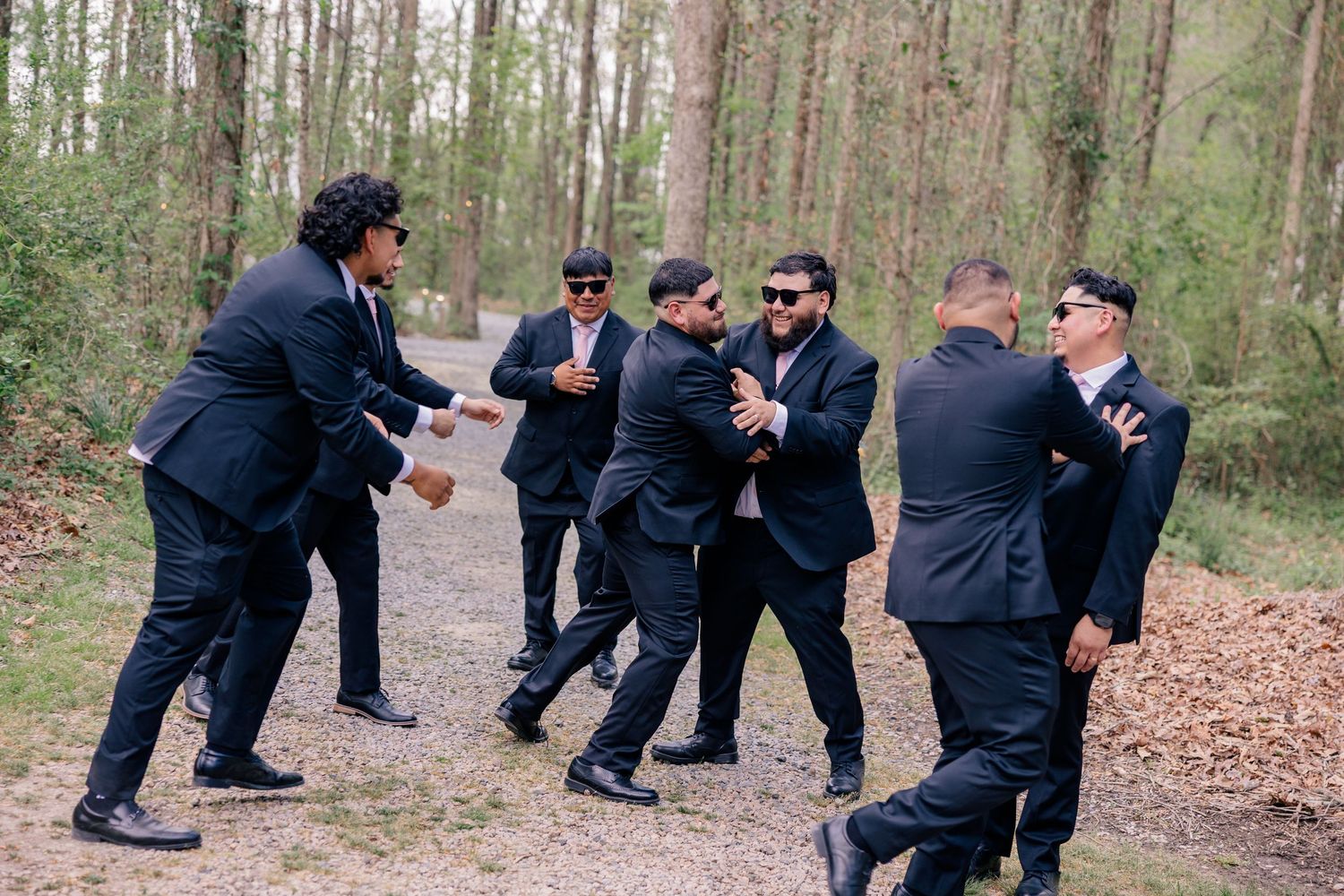 Groomsmen in black suits pose playfully in a wooded outdoor setting.