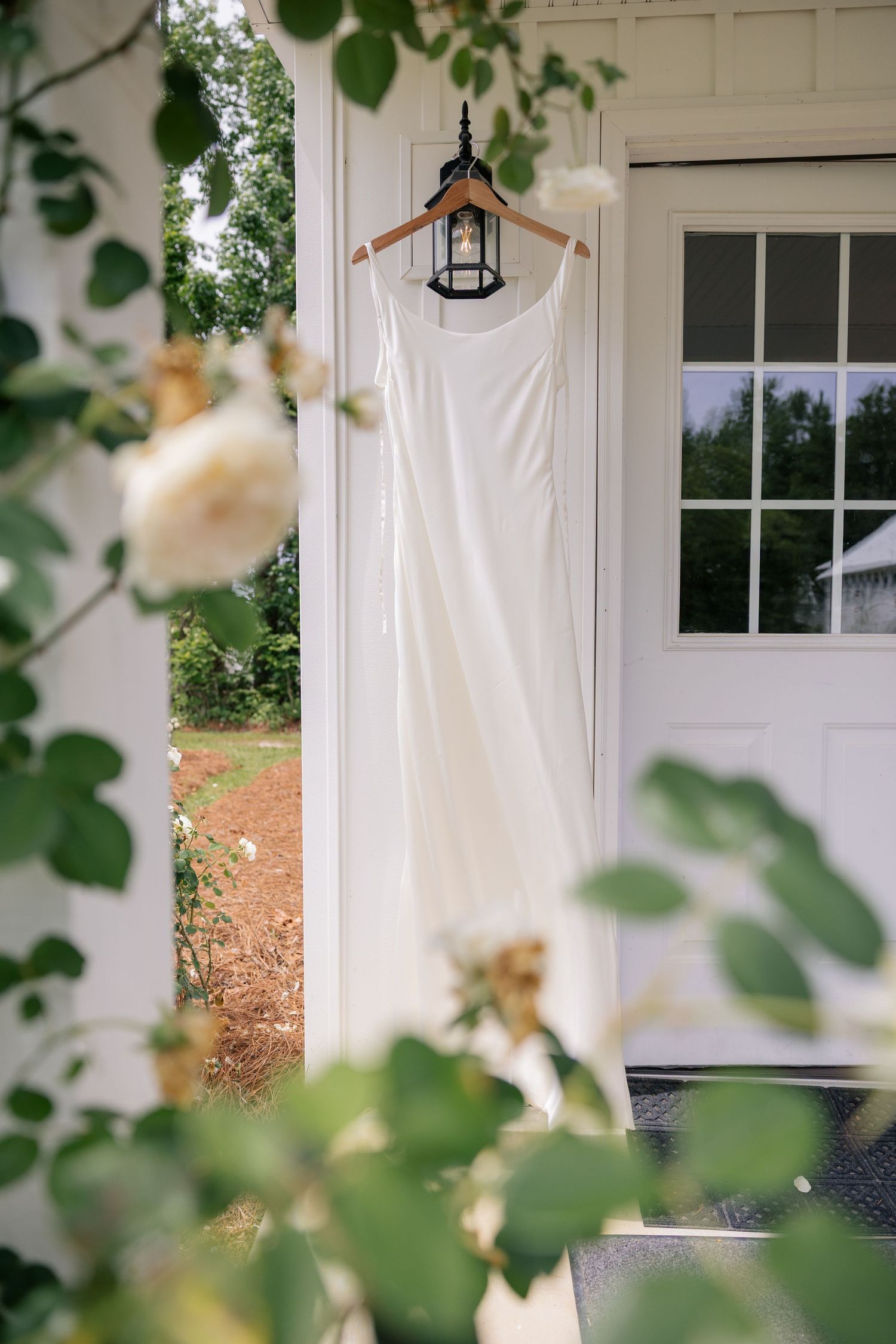 White curtains frame windows on a rustic white chapel exterior with green foliage peeking into view.