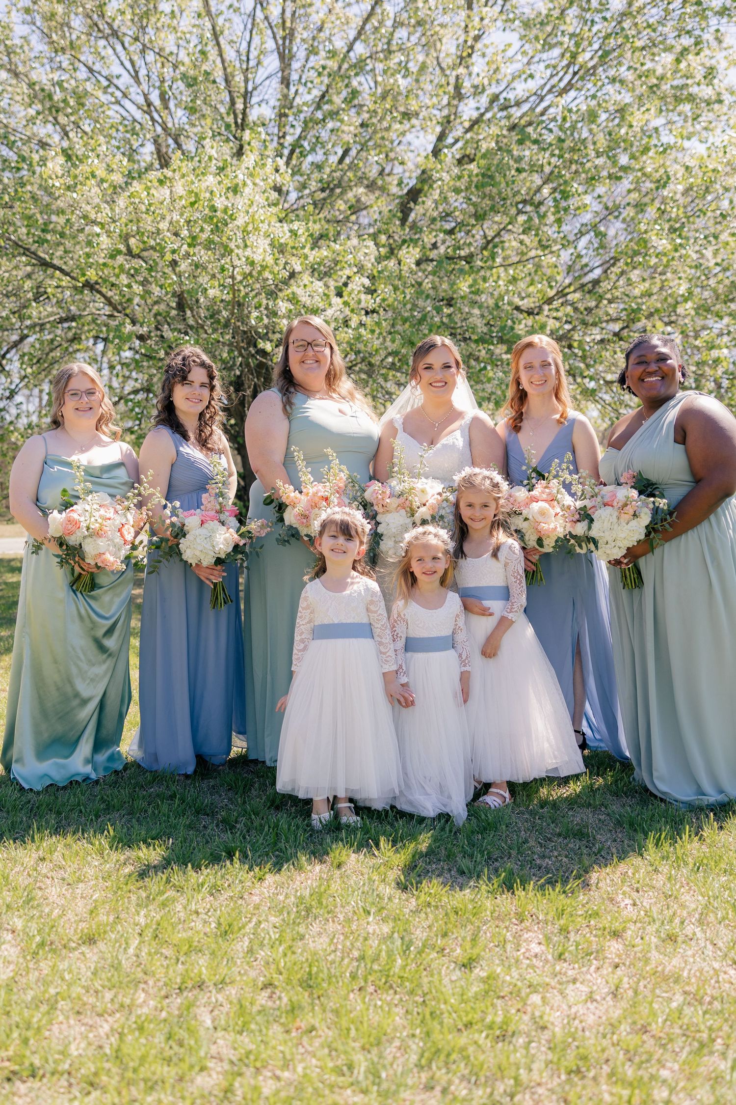 Wedding party group poses together on sunny grass field wearing mix of sage green and blue dresses.