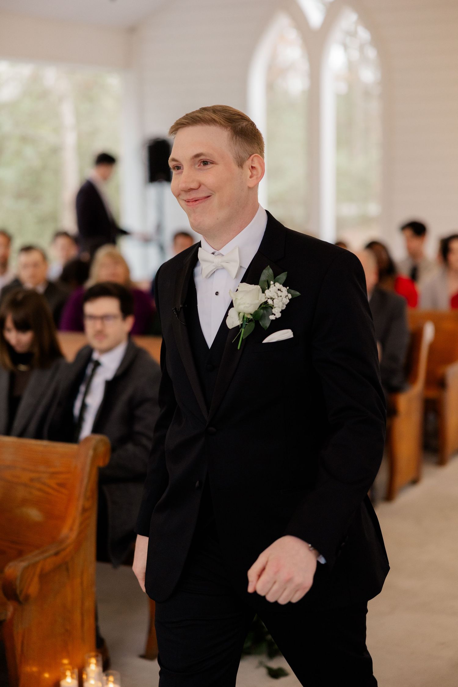 Groomsmen in black tuxedos share a laugh while seated in wooden church pews.