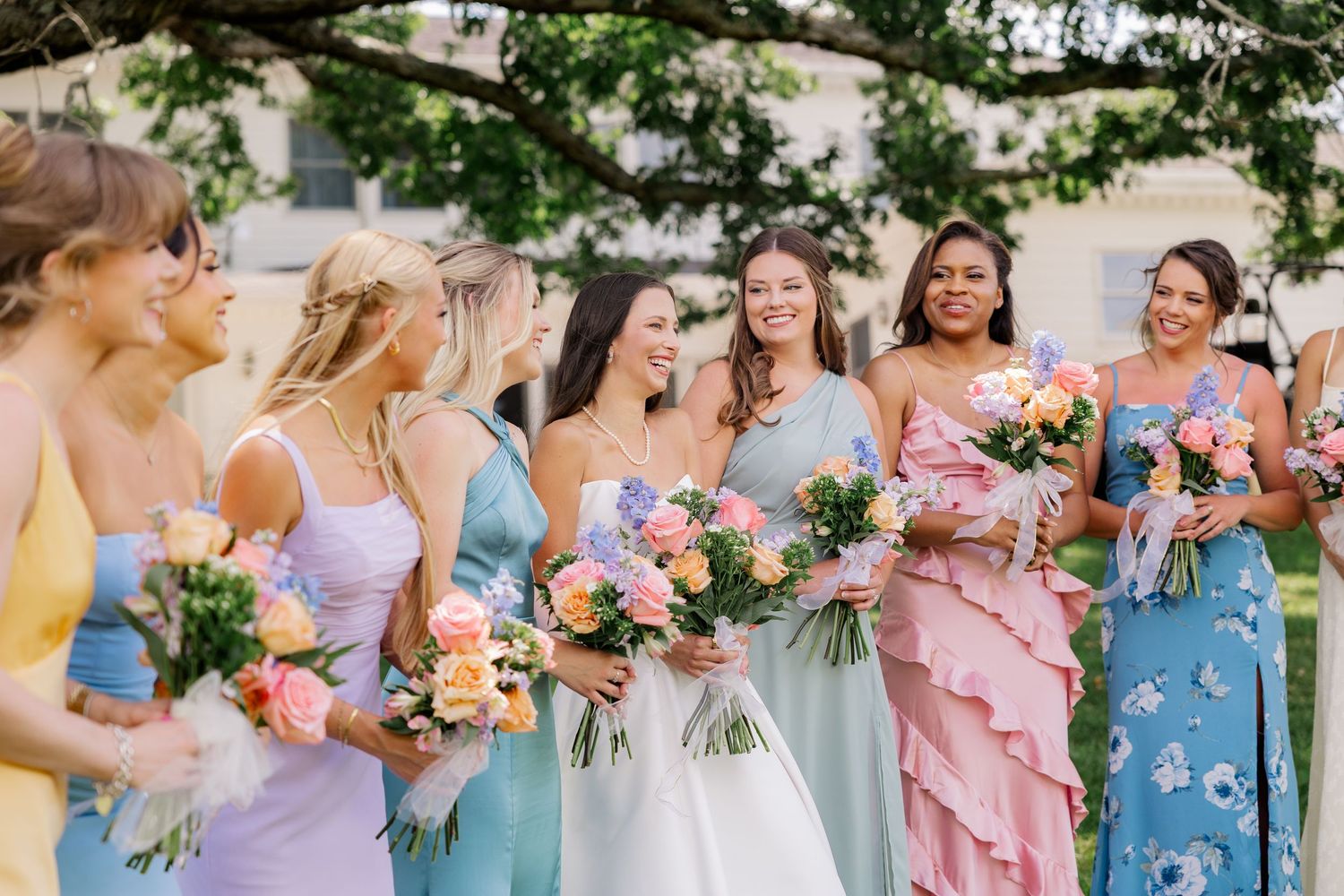 Bridesmaids in pastel blue and purple dresses gather outdoors holding colorful bouquets.