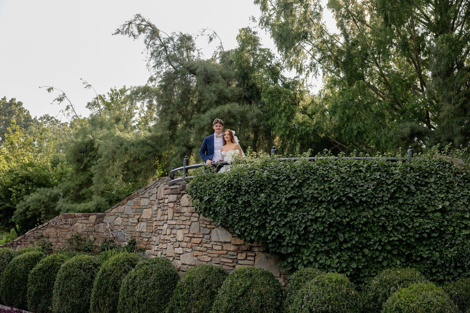 A couple shares a romantic moment atop a stone wall in a manicured garden.