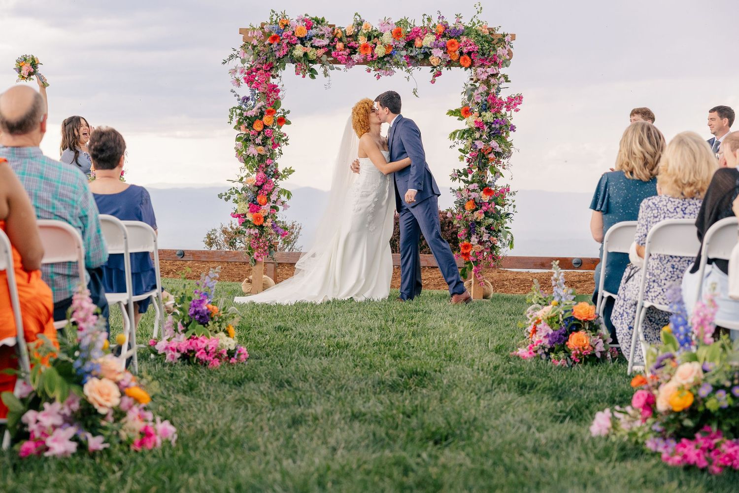 Bride and groom kissing at outdoor wedding ceremony under colorful floral arch with ocean view.