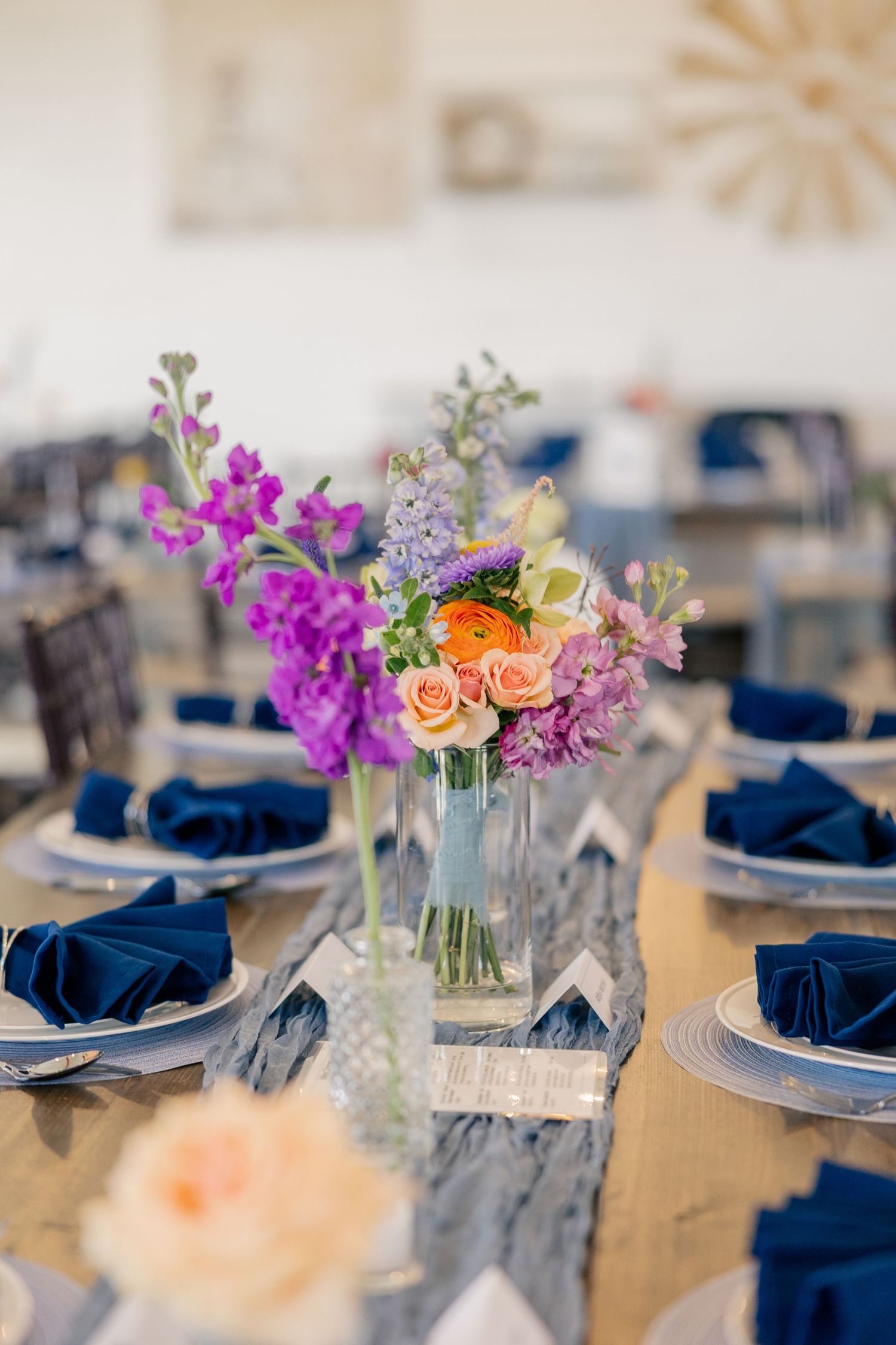 Elegant wedding reception table with colorful floral centerpiece and navy blue napkins on wooden tables.