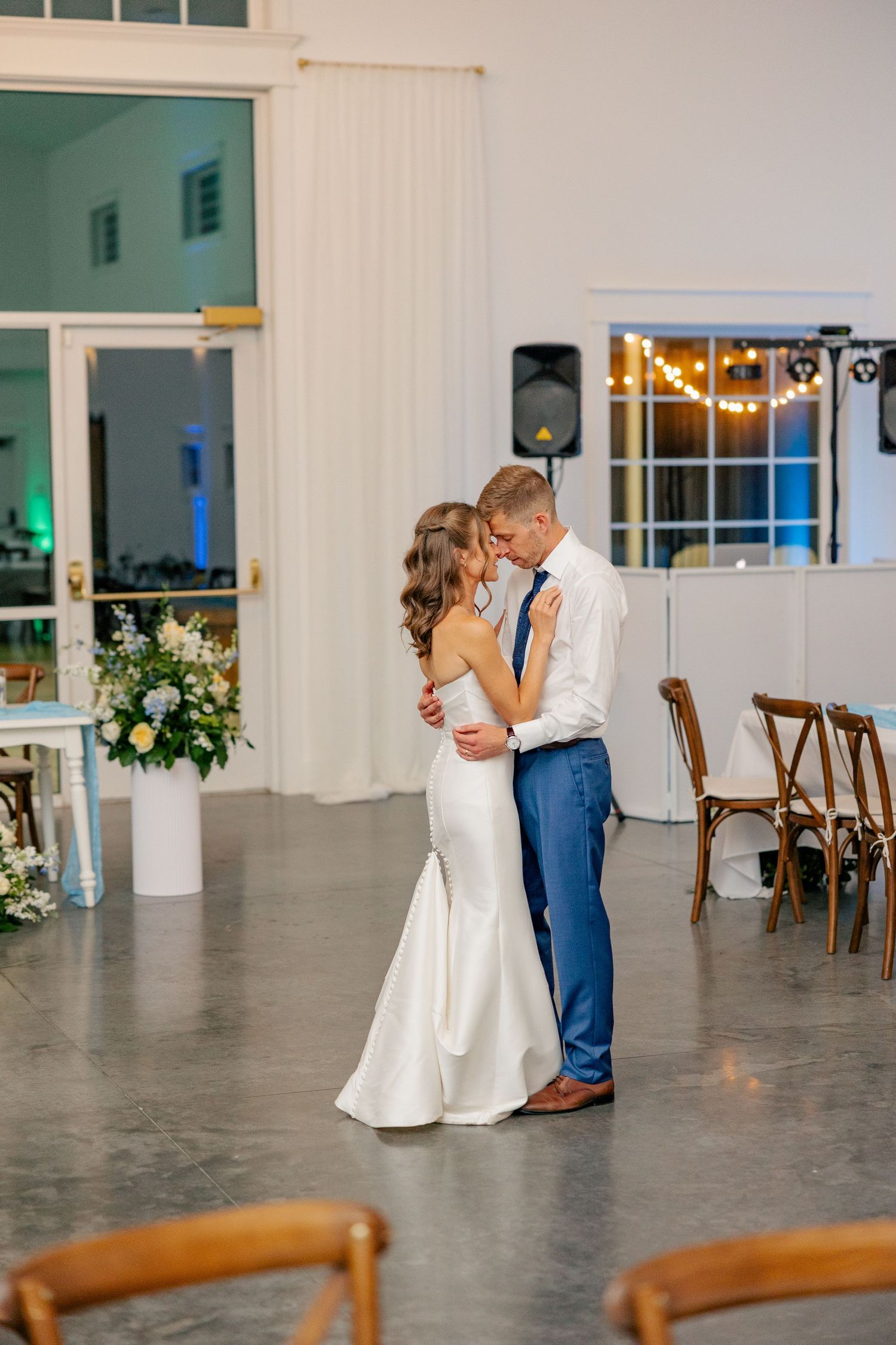 Bride and groom share intimate first dance embrace in elegant white reception hall with string lights.
