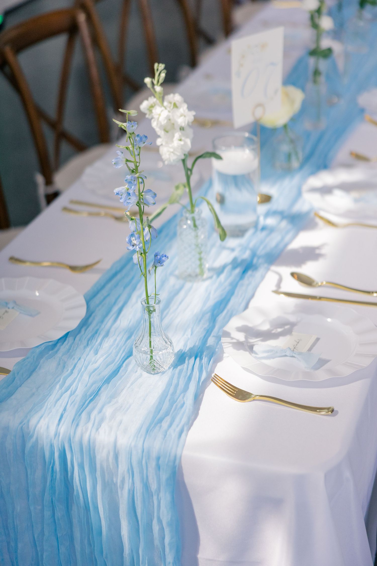 Elegant wedding table with light blue gauze runner, gold cutlery, and delicate floral stems.
