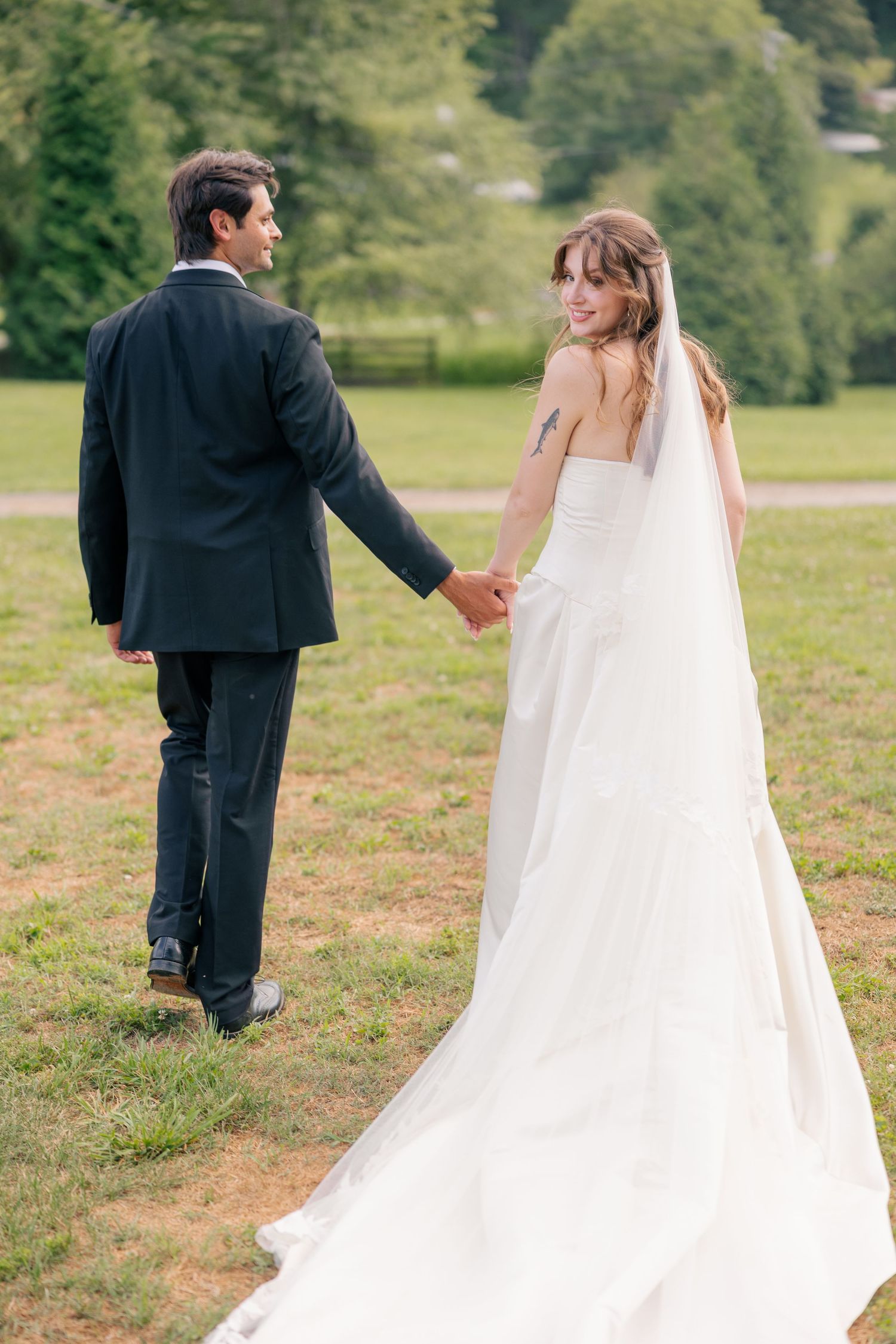Bride in white gown with veil glances back while holding hands with groom walking across a green outdoor field.