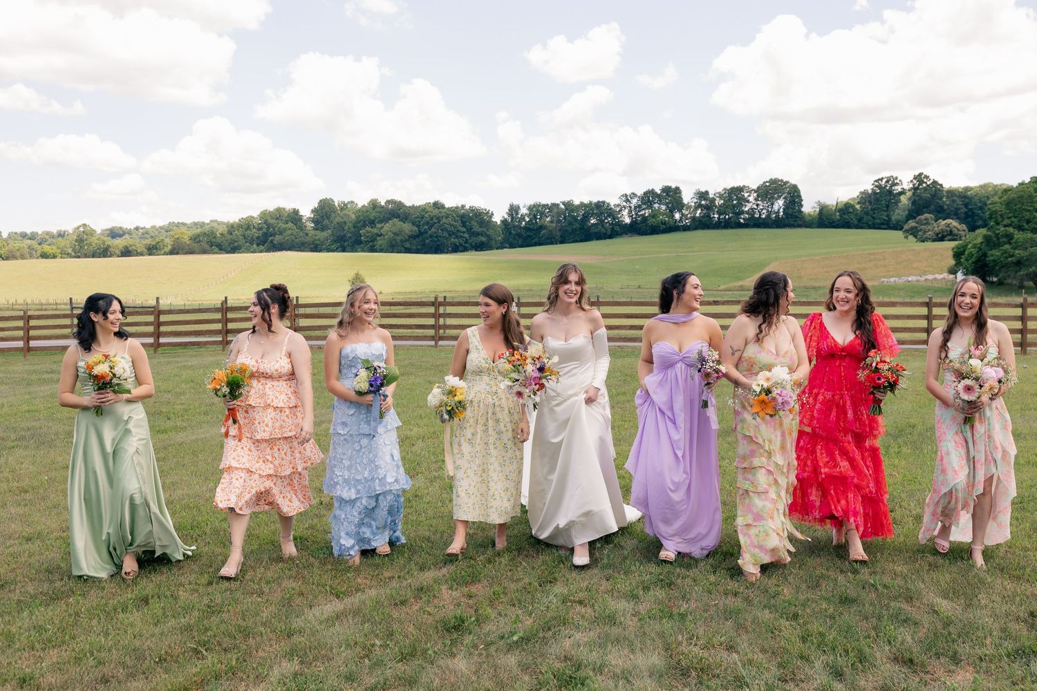 Bride walks with bridesmaids in mismatched colorful gowns across green farm field on wedding day.