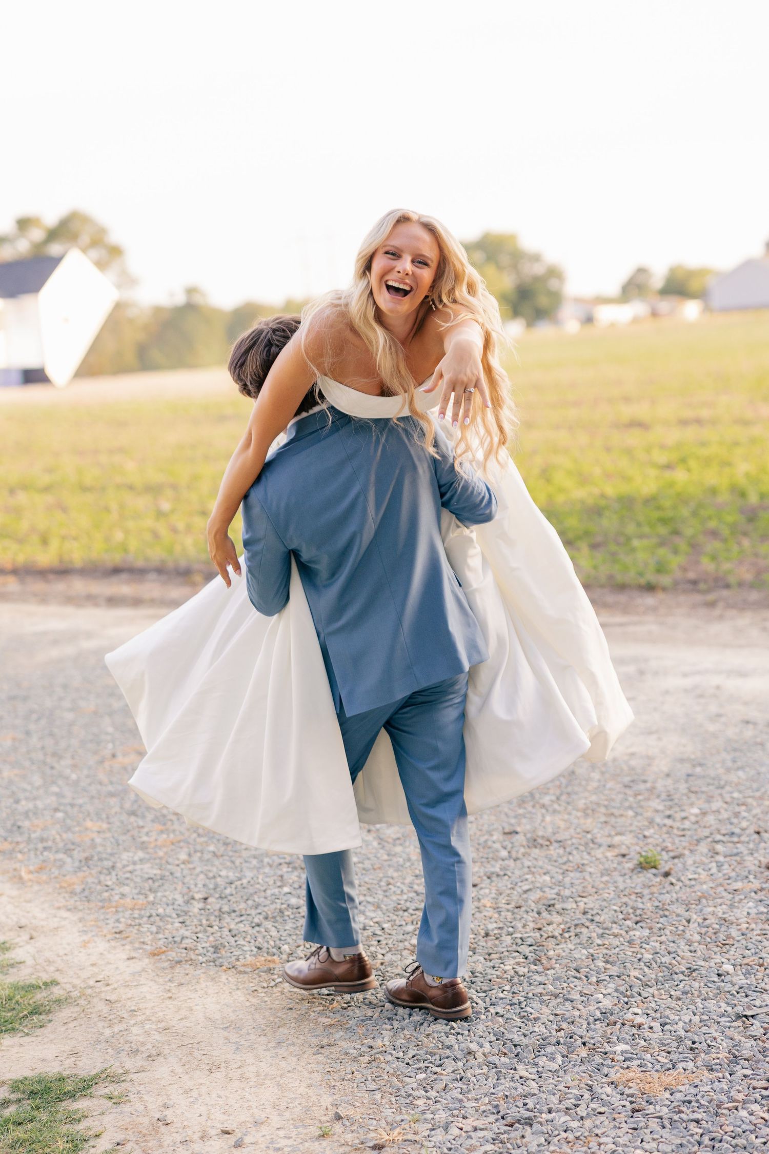 Joyful bride laughing as groom carries her on his back on a gravel path at an outdoor farm wedding venue.
