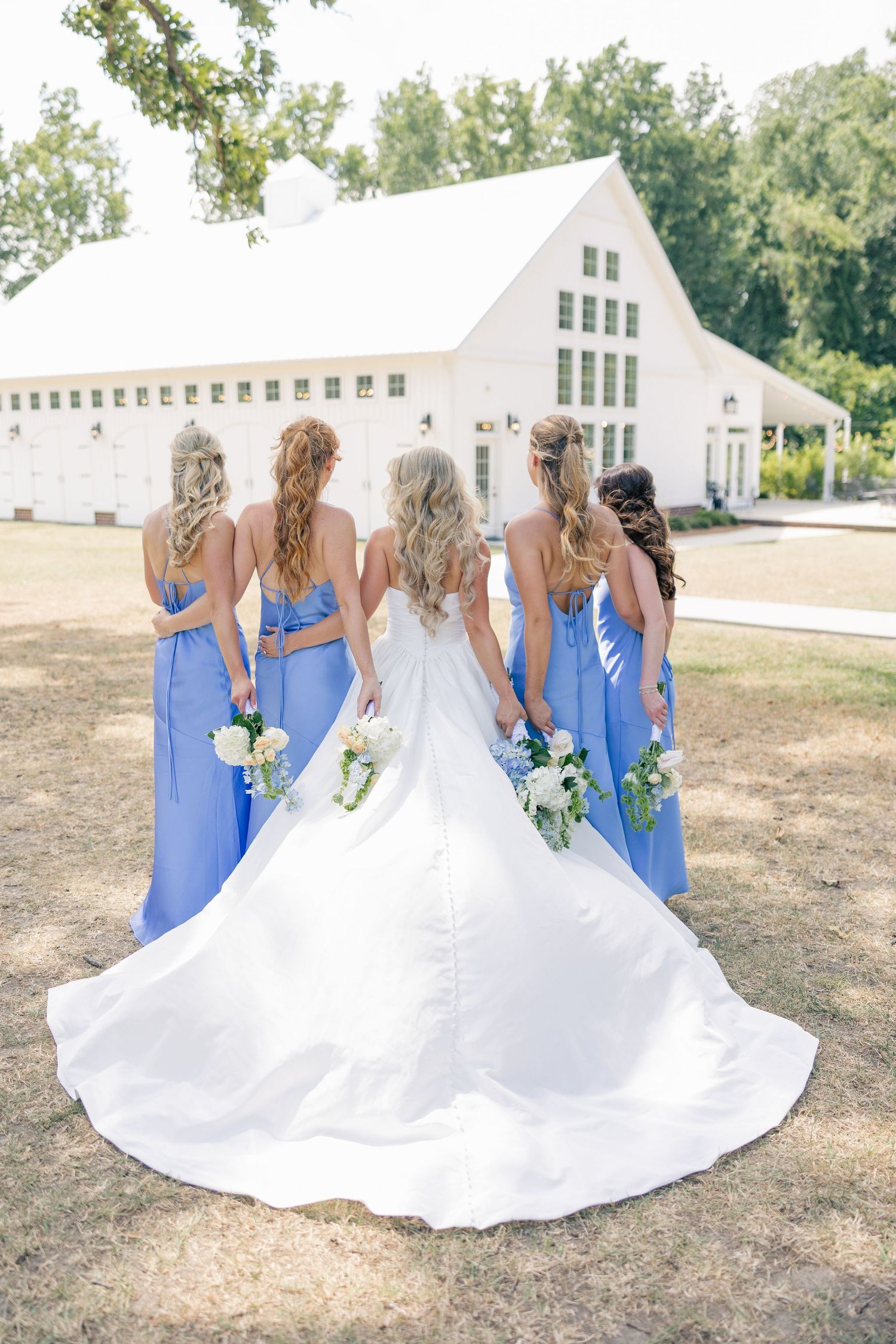 Bride and four bridesmaids in blue dresses stand facing white barn venue holding bouquets.