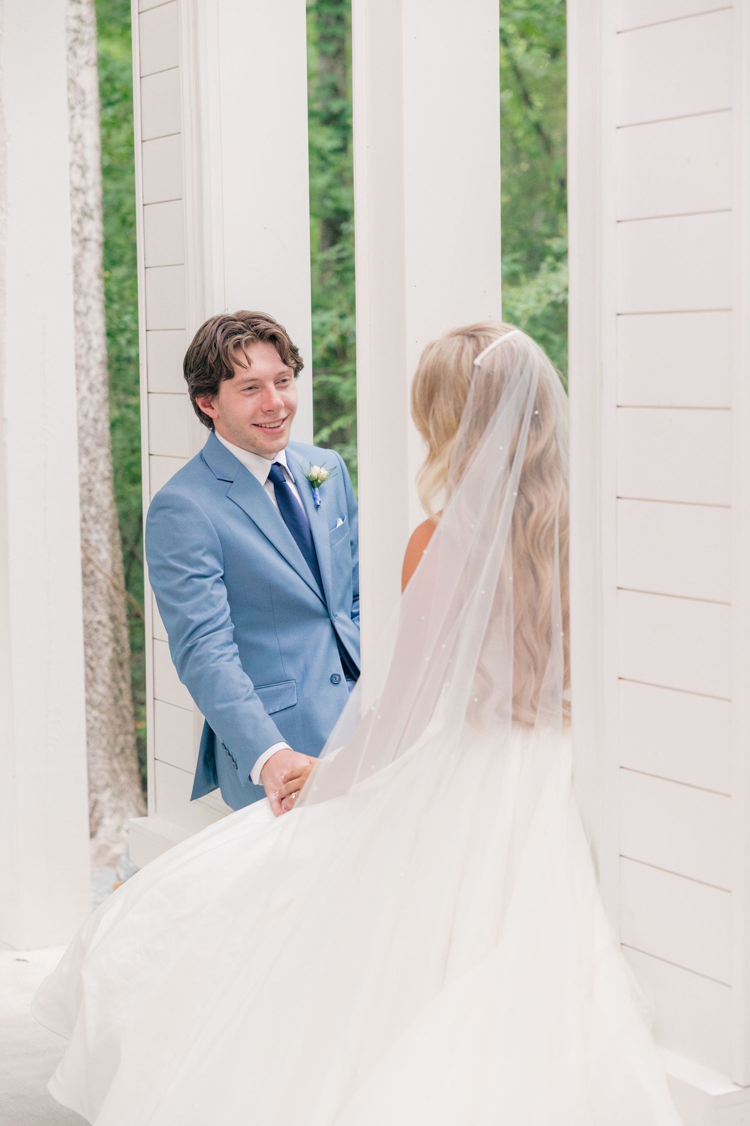 Smiling groom in light blue suit holds hands with veiled bride between white columns.