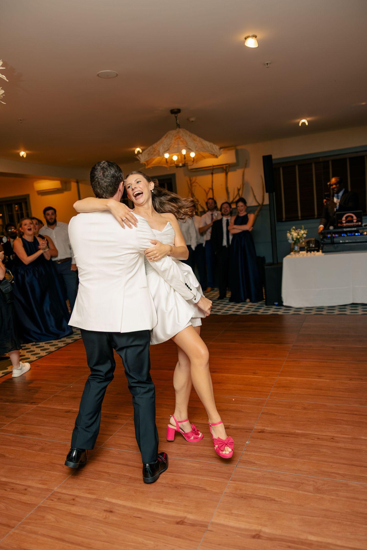 Joyful bride in short white dress and pink heels dancing with groom during first dance at reception.