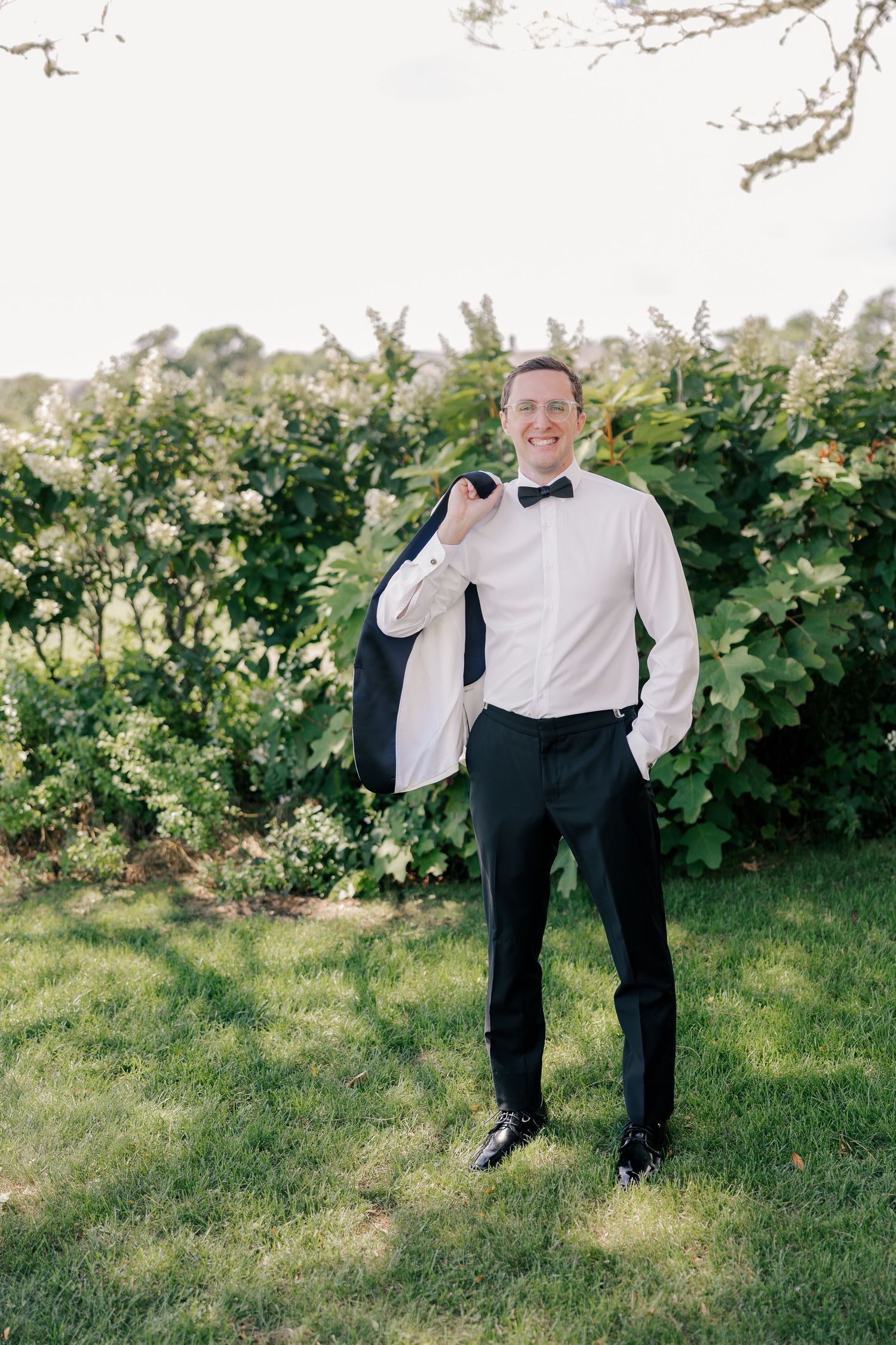 Dapper groom in a white dress shirt and black bow tie holds his tuxedo jacket over his shoulder outdoors.