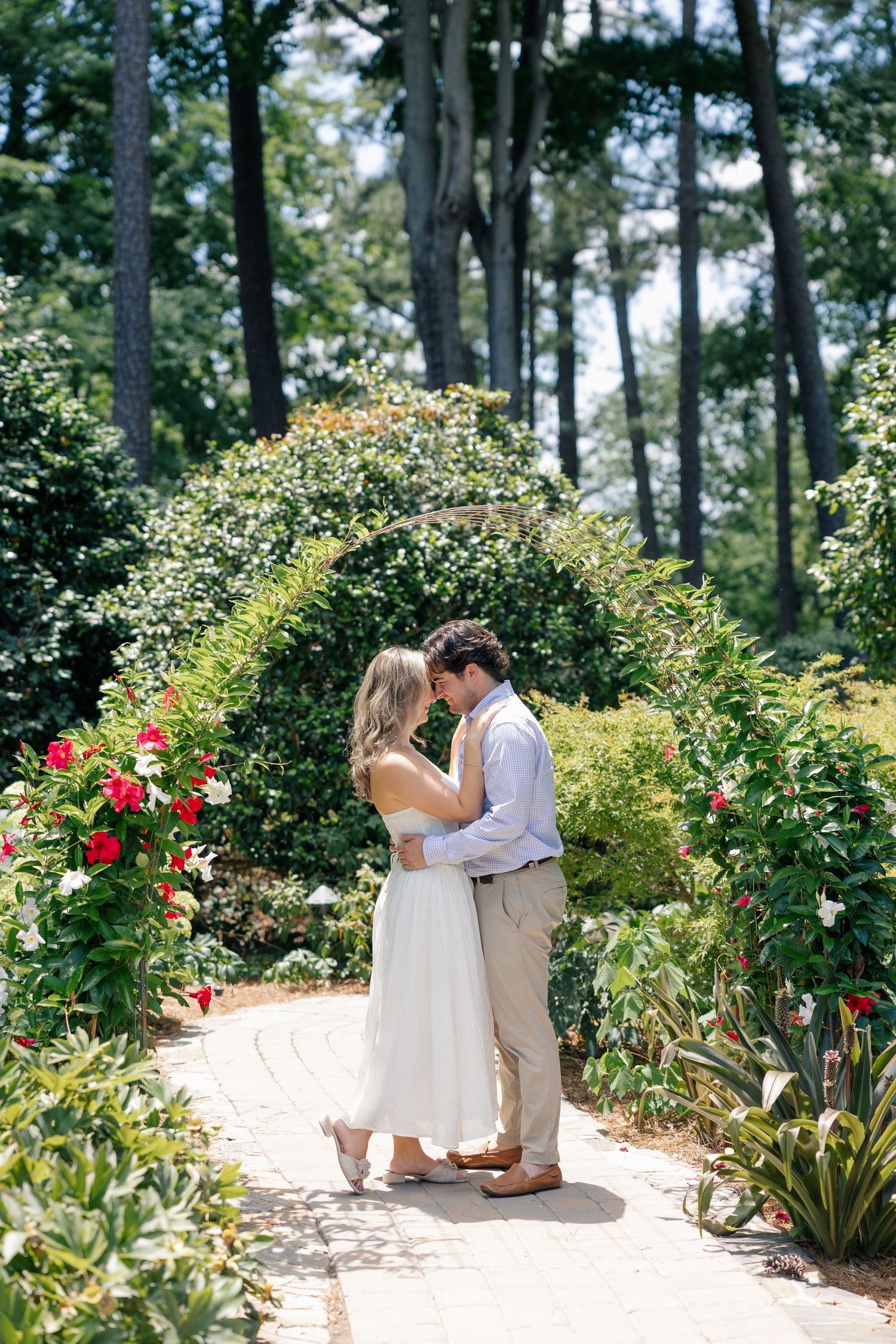 Engaged couple sharing a tender moment on a garden path surrounded by lush greenery and red flowers.