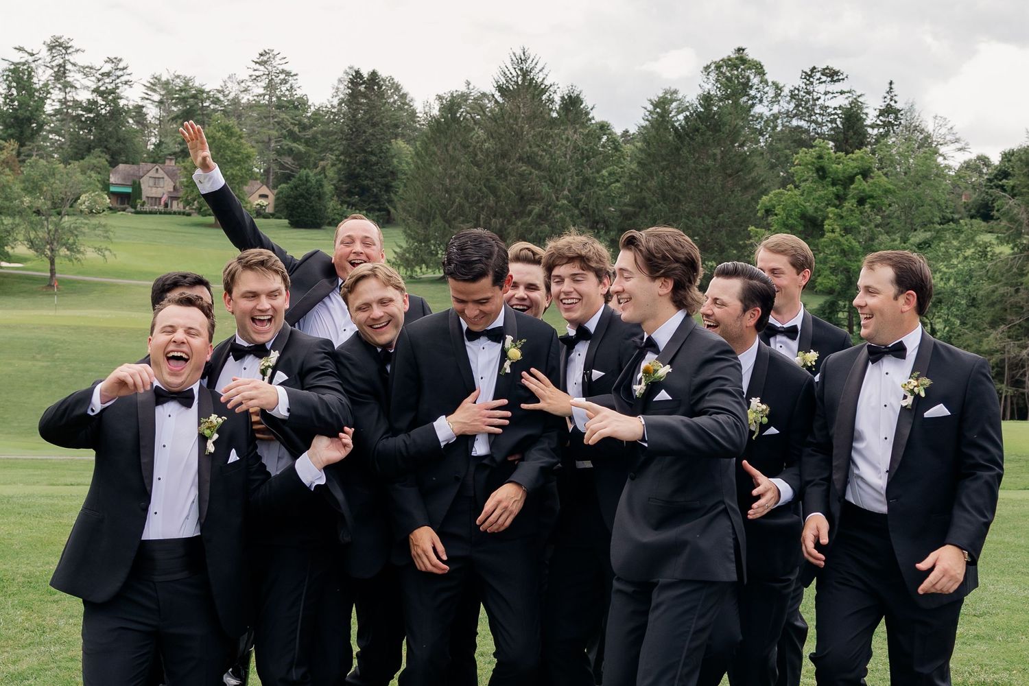 Groomsmen in black tuxedos laughing and celebrating outdoors on a golf course at a wedding.