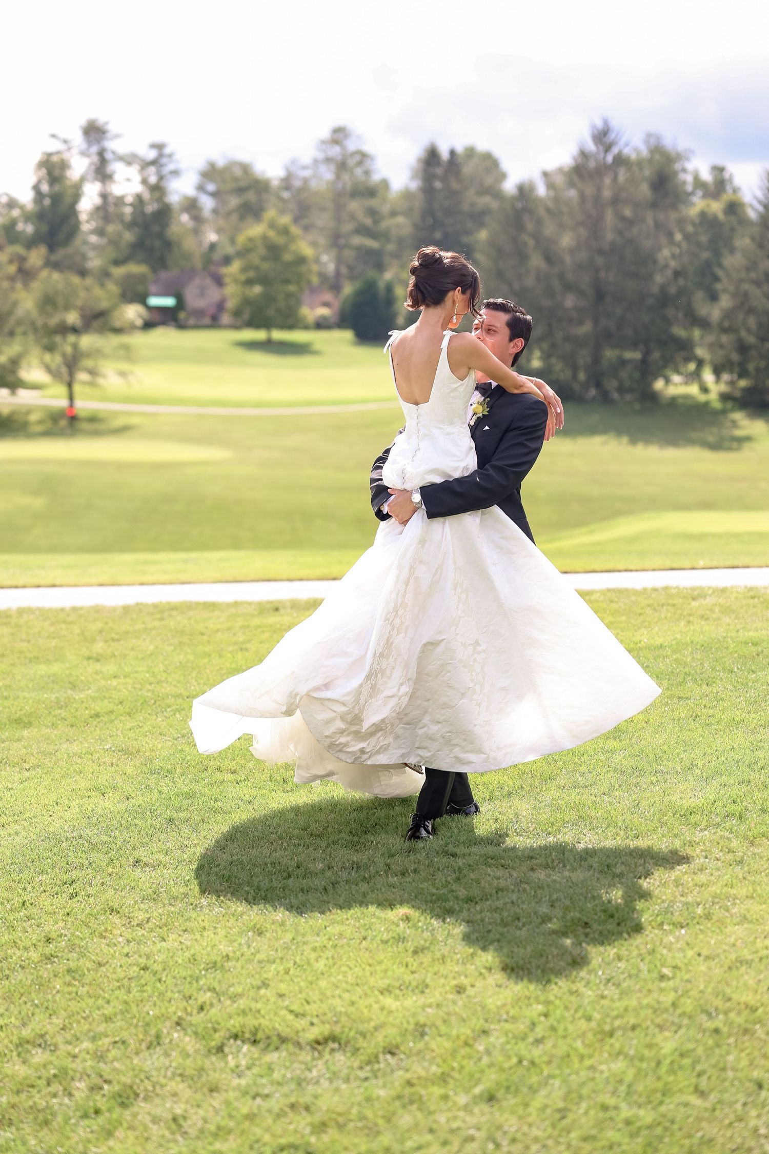 Groom lifting and spinning bride in white gown on lush green lawn during outdoor wedding celebration.