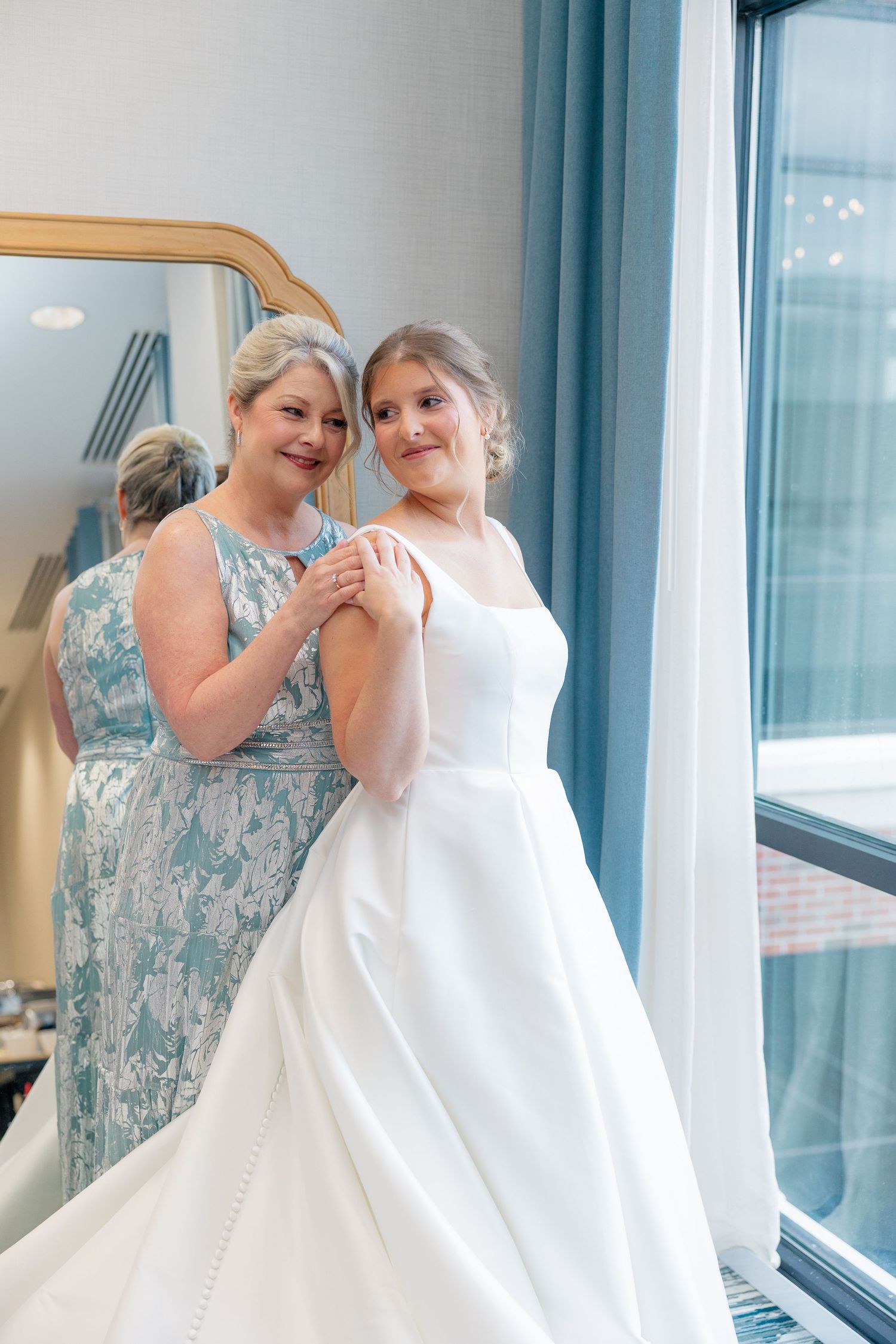 Mother helps bride in sleek white ball gown prepare near bright window before wedding ceremony.