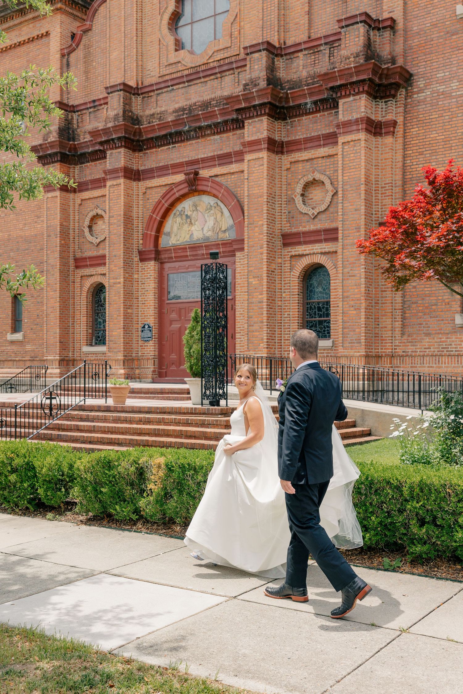 Bride and groom walk hand in hand outside historic red brick Basilica Shrine of St. Mary church.