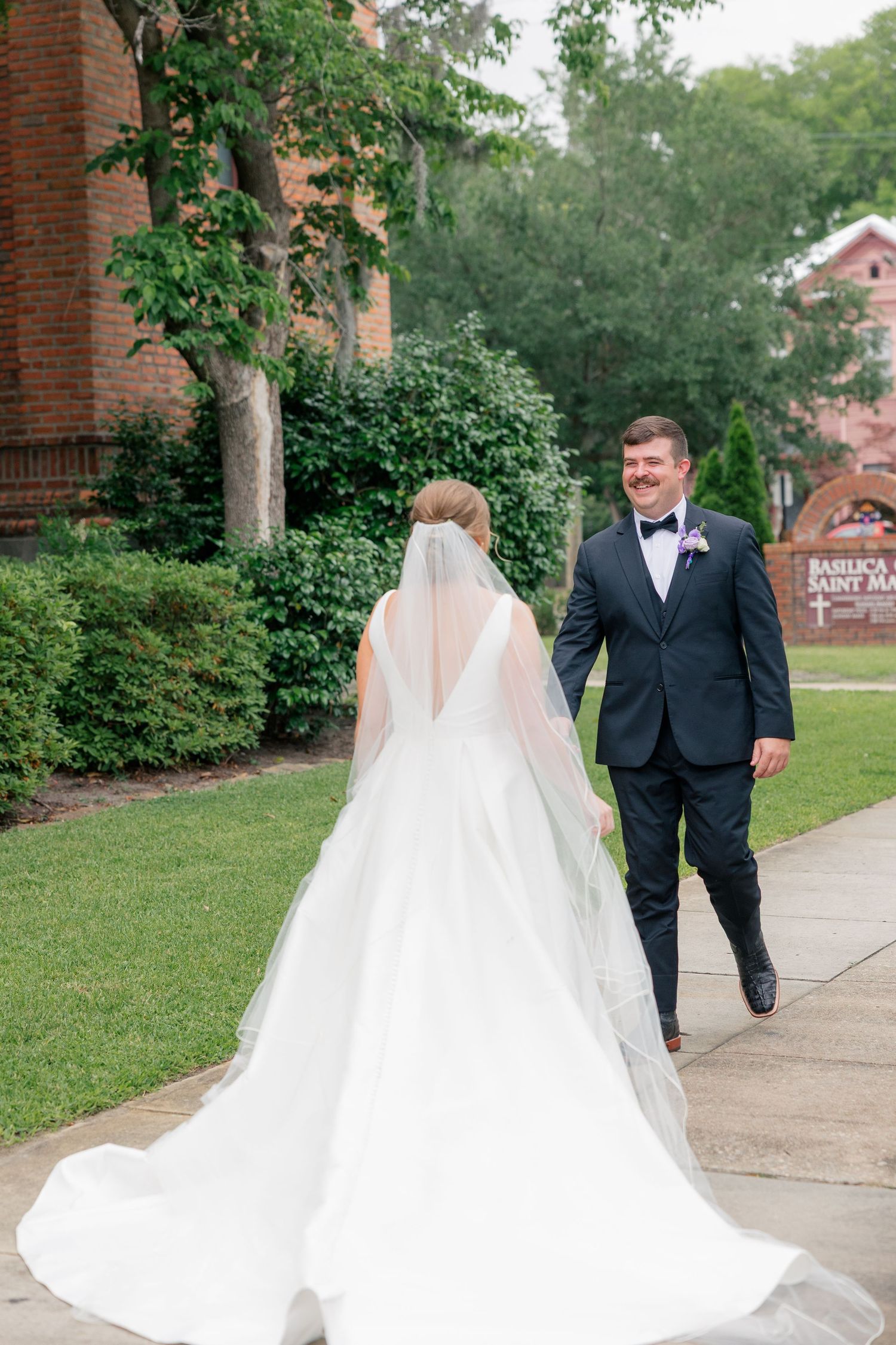 Bride in white gown with cathedral veil walking with groom in black tuxedo outside church.