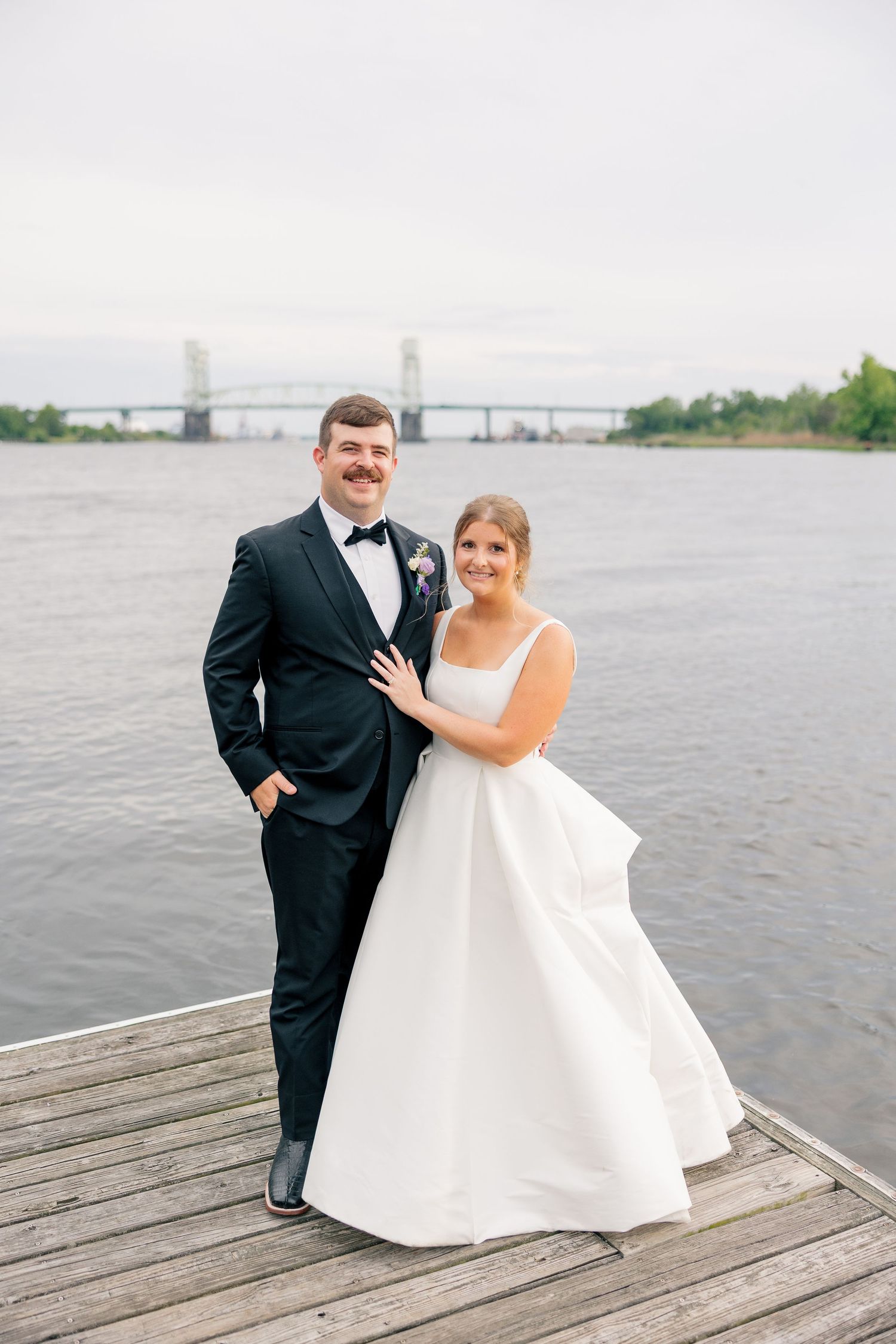 Bride and groom posing on a wooden dock by the water with a bridge in the background on their wedding day.