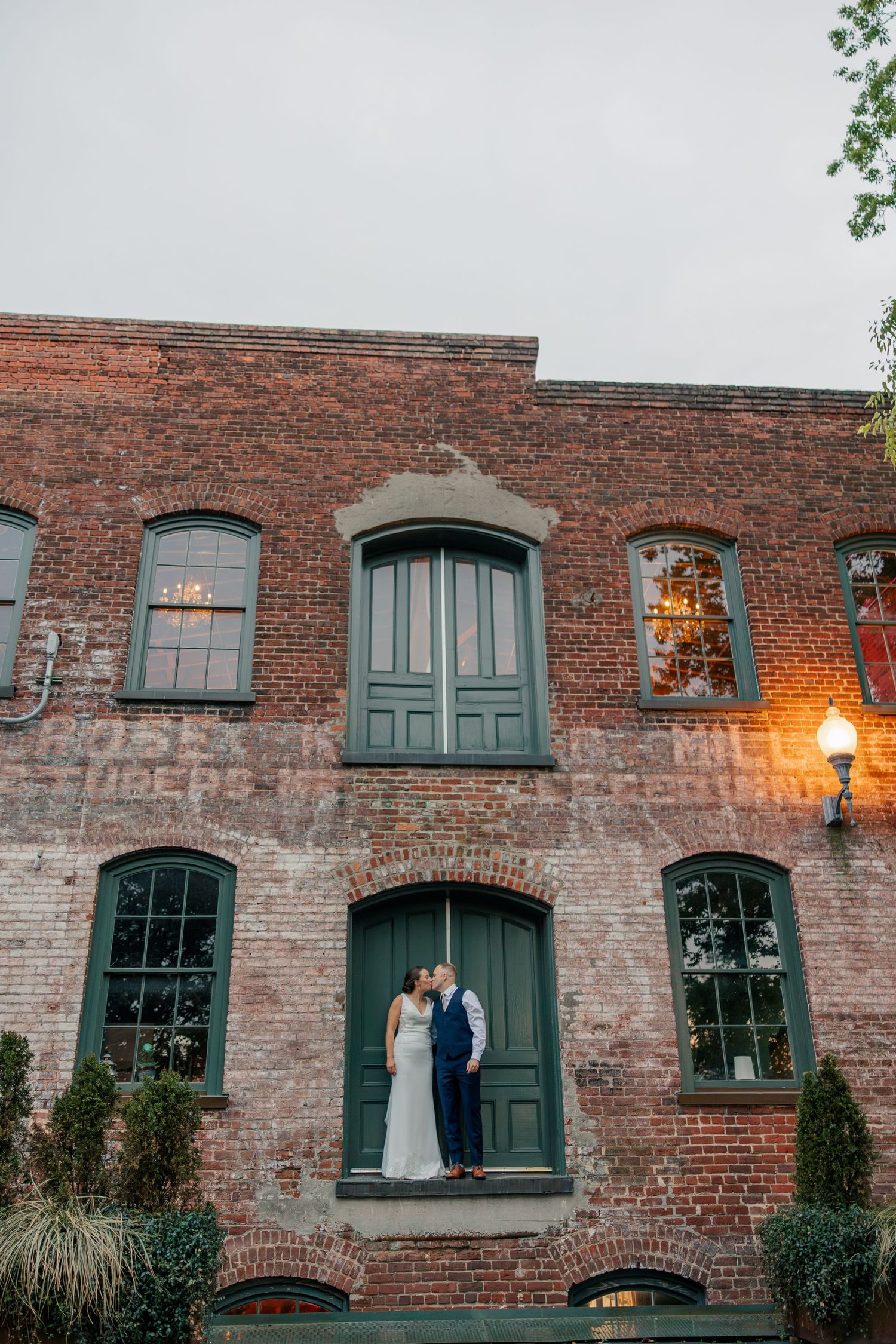 Bride and groom share romantic kiss in doorway of historic red brick building at dusk.