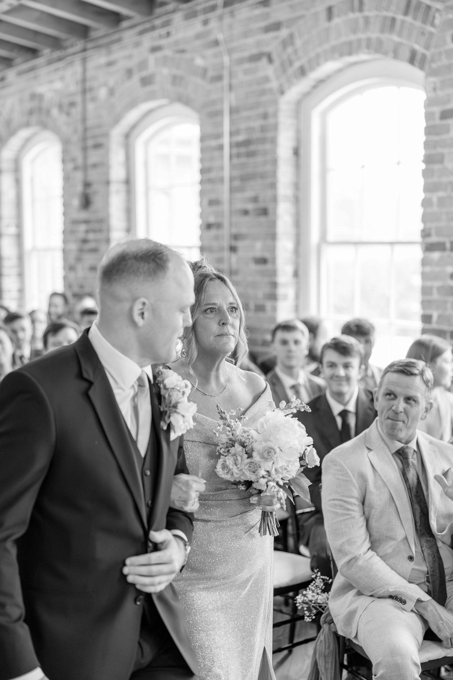 Bride walks down the aisle holding a bouquet in a sparkly gown during an indoor wedding ceremony in a brick venue.