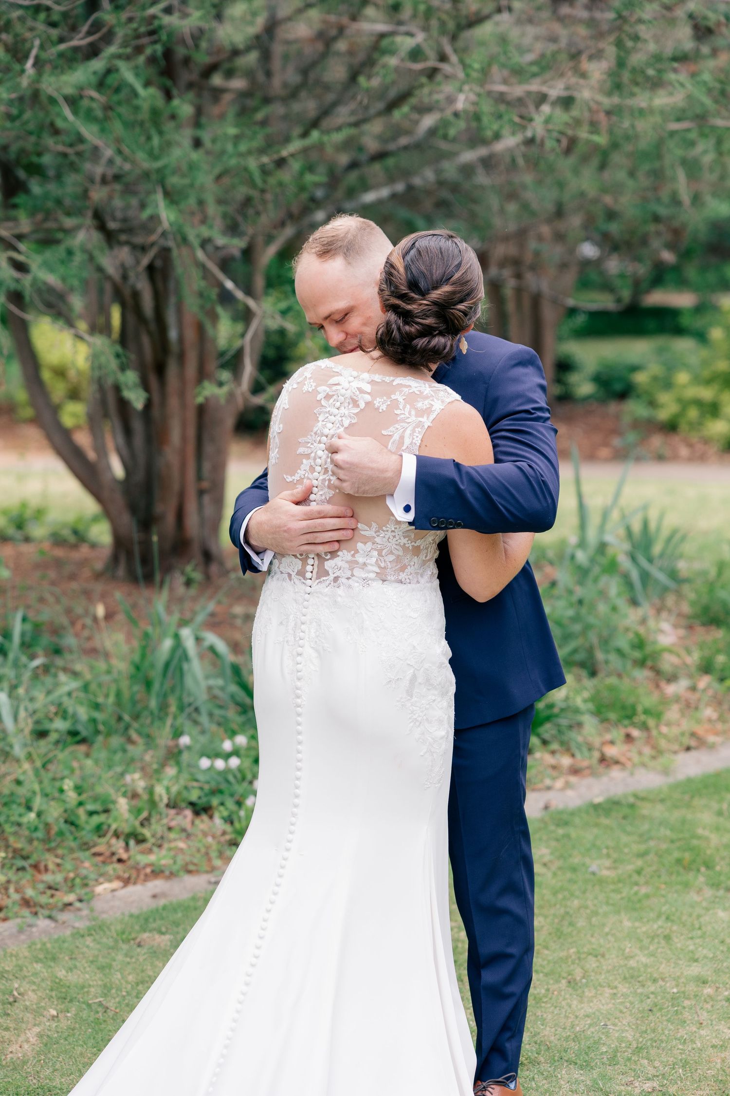 Bride and groom share an emotional first look embrace outdoors, her lace back wedding gown on display.