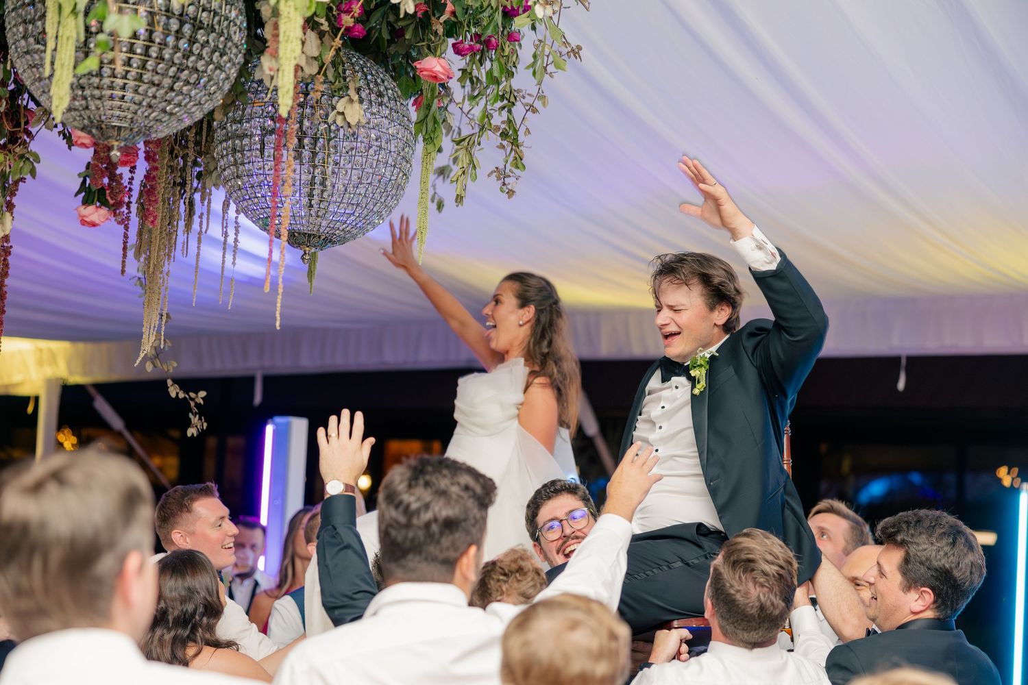 Bride and groom lifted by guests during joyful wedding reception celebration under floral chandelier.