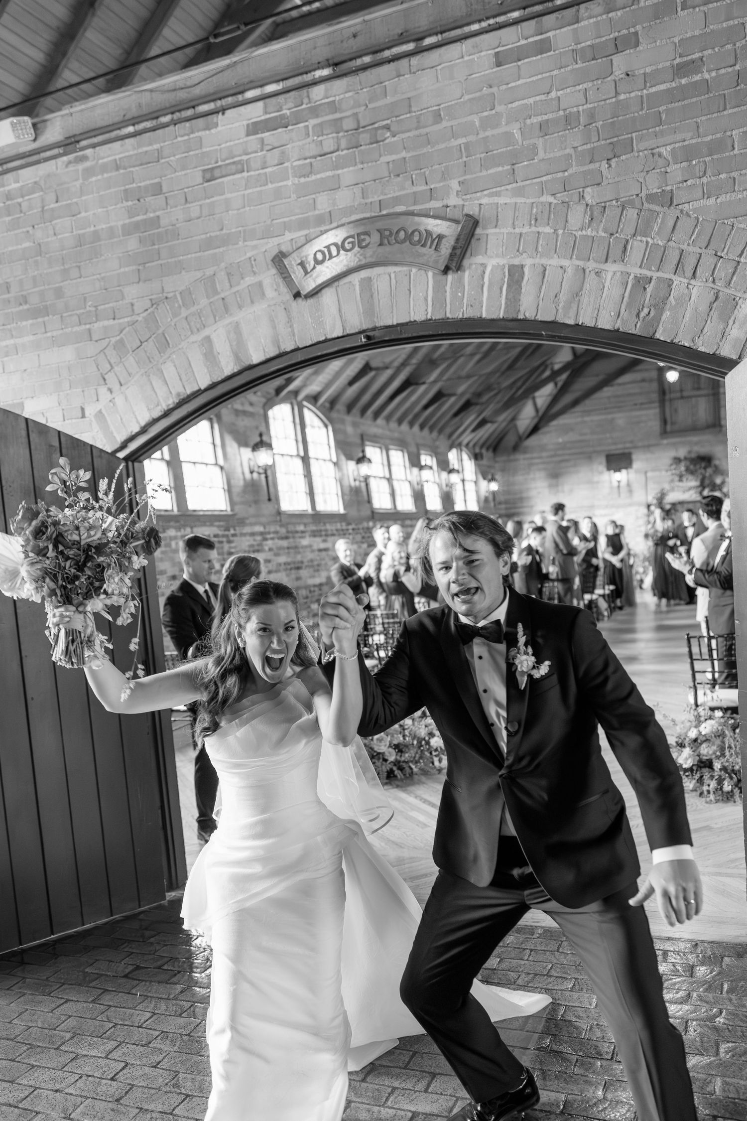 Joyful bride and groom celebrate their grand entrance through an arched brick doorway in black and white photo.