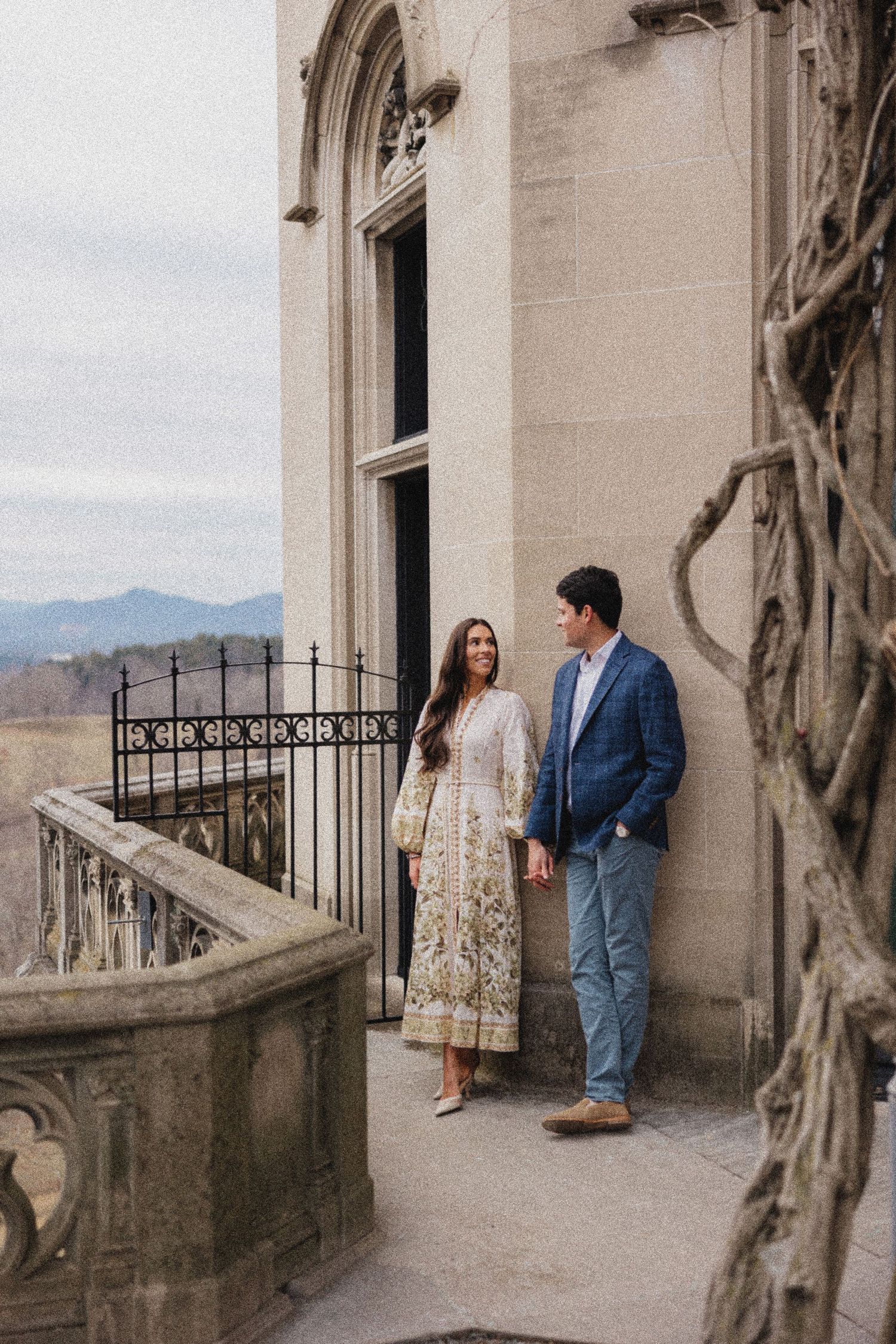 A couple stands on an ornate stone balcony of a historic Gothic-style building with mountains visible in the distance.