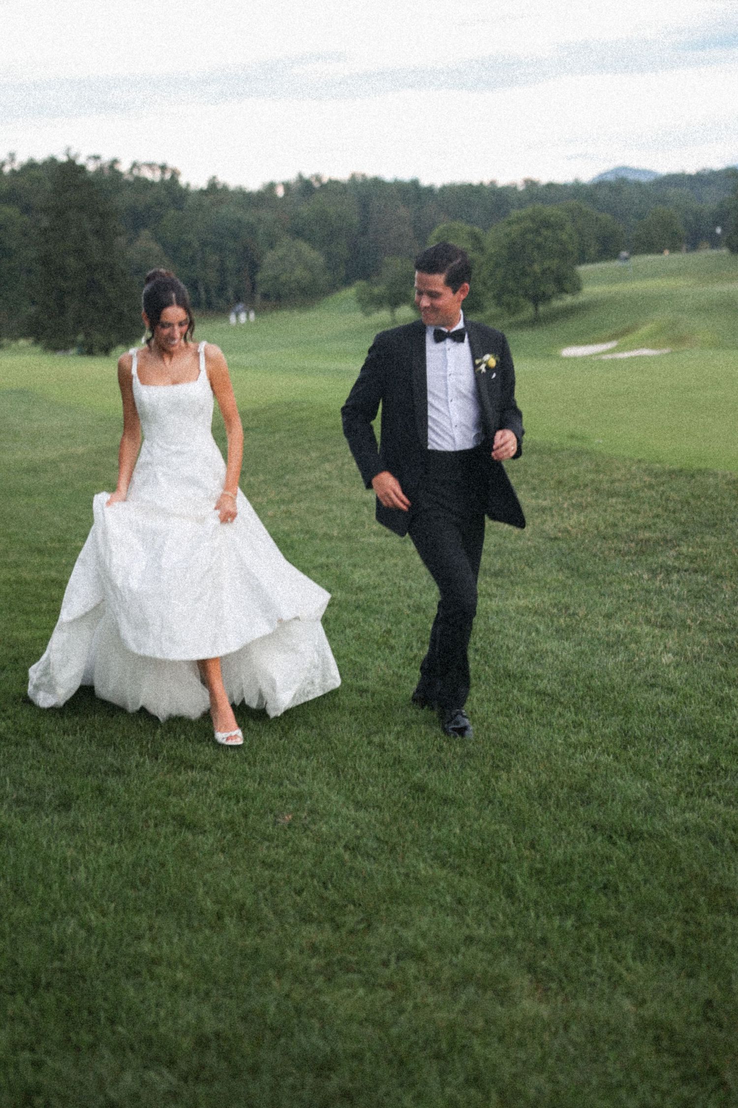Bride in white gown and groom in black tuxedo walking together on a lush green golf course.