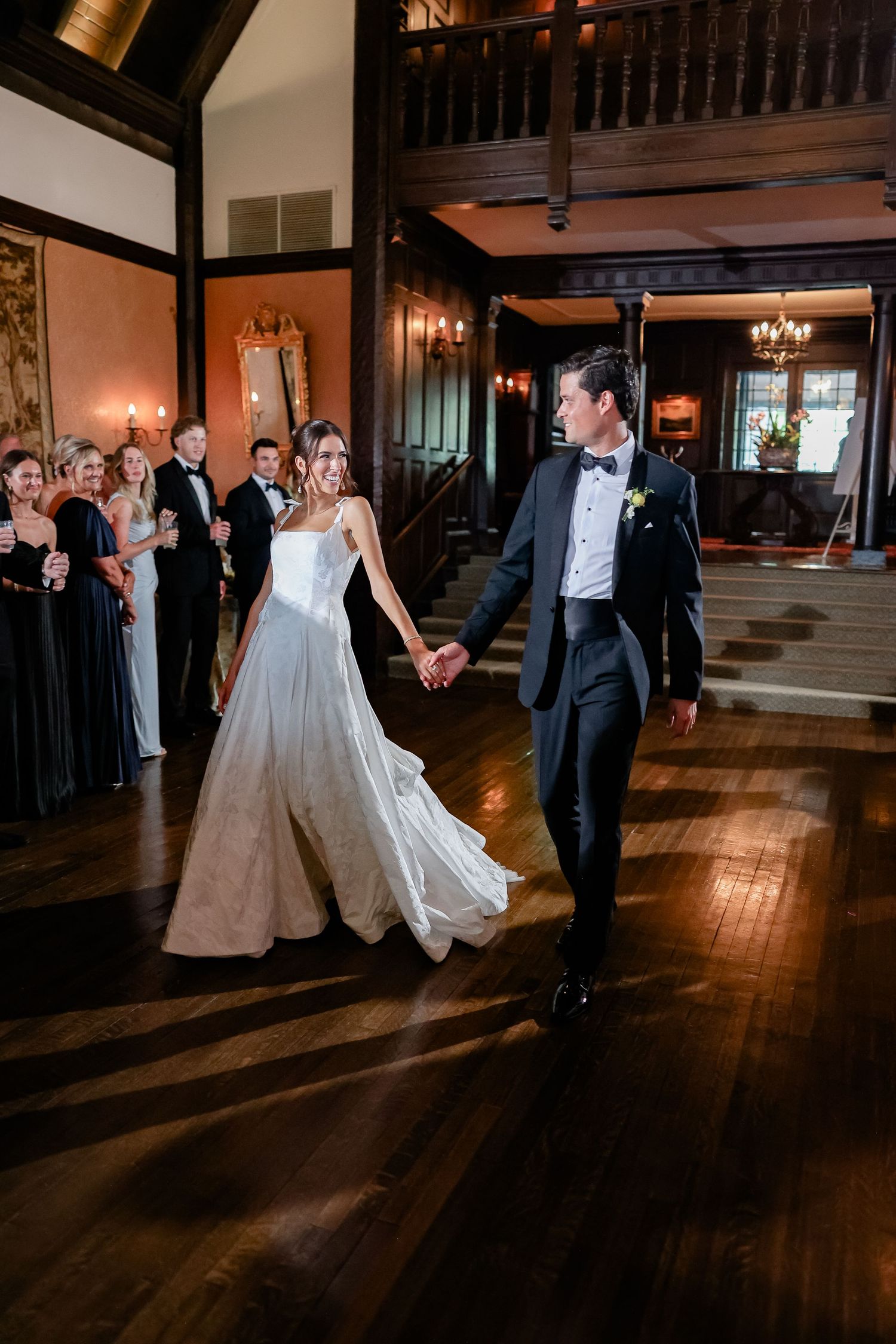 Bride and groom hold hands walking onto dance floor at elegant wedding reception with guests watching.