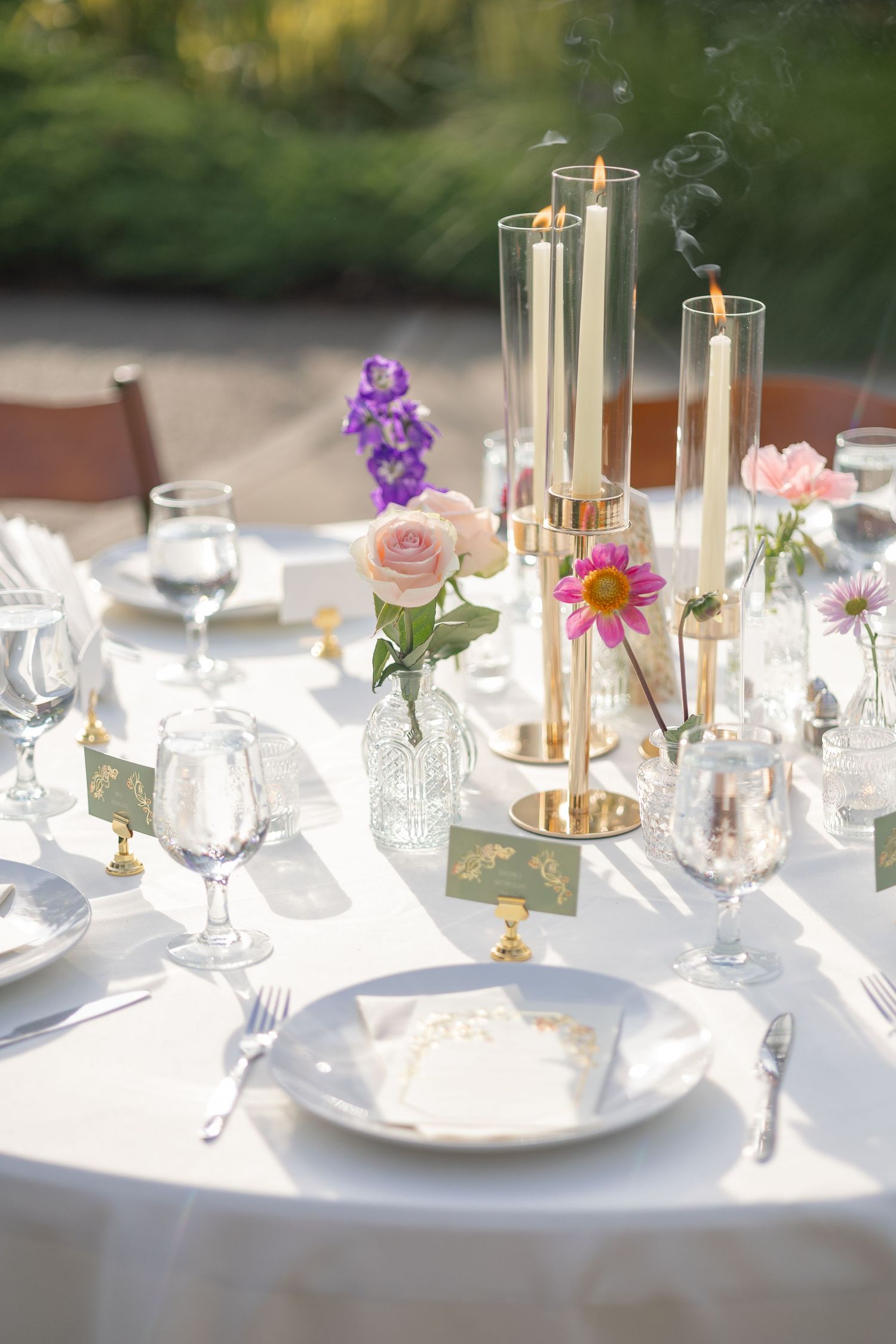 Wedding table settings with delicate pink and purple flowers in glass vases on white tablecloths.