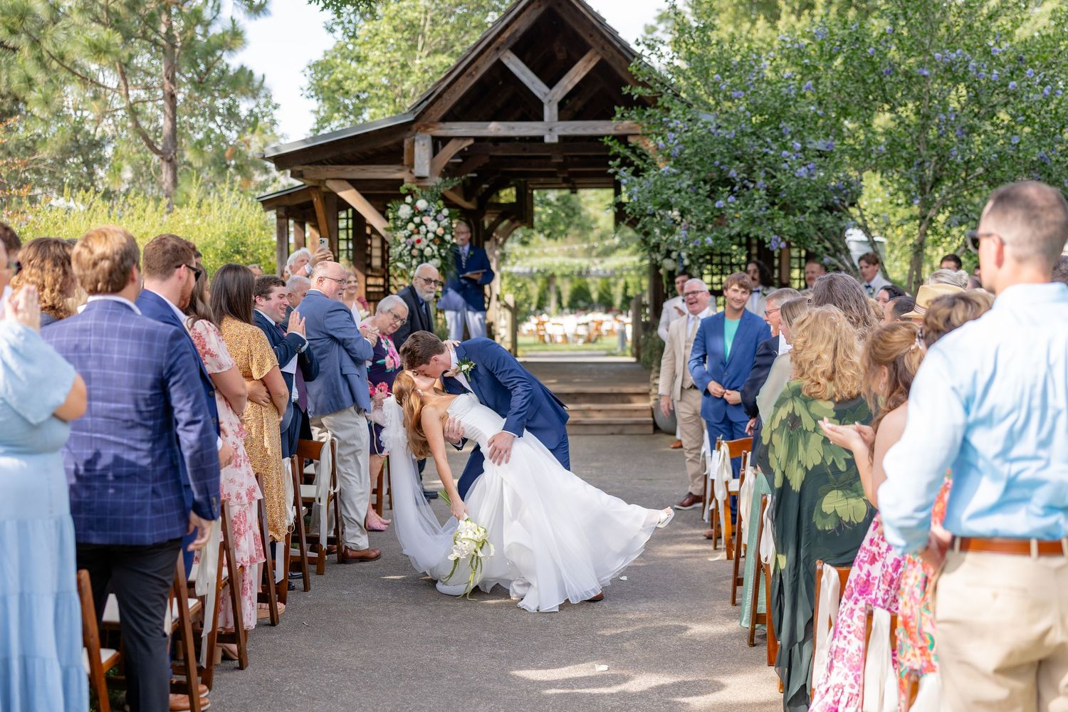 Wide view of garden wedding ceremony with guests seated beneath wooden pavilion structure.