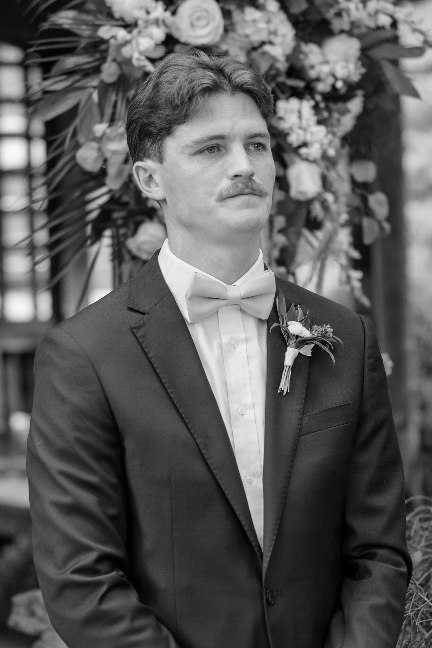 Black and white portrait of groom in formal attire with floral backdrop.