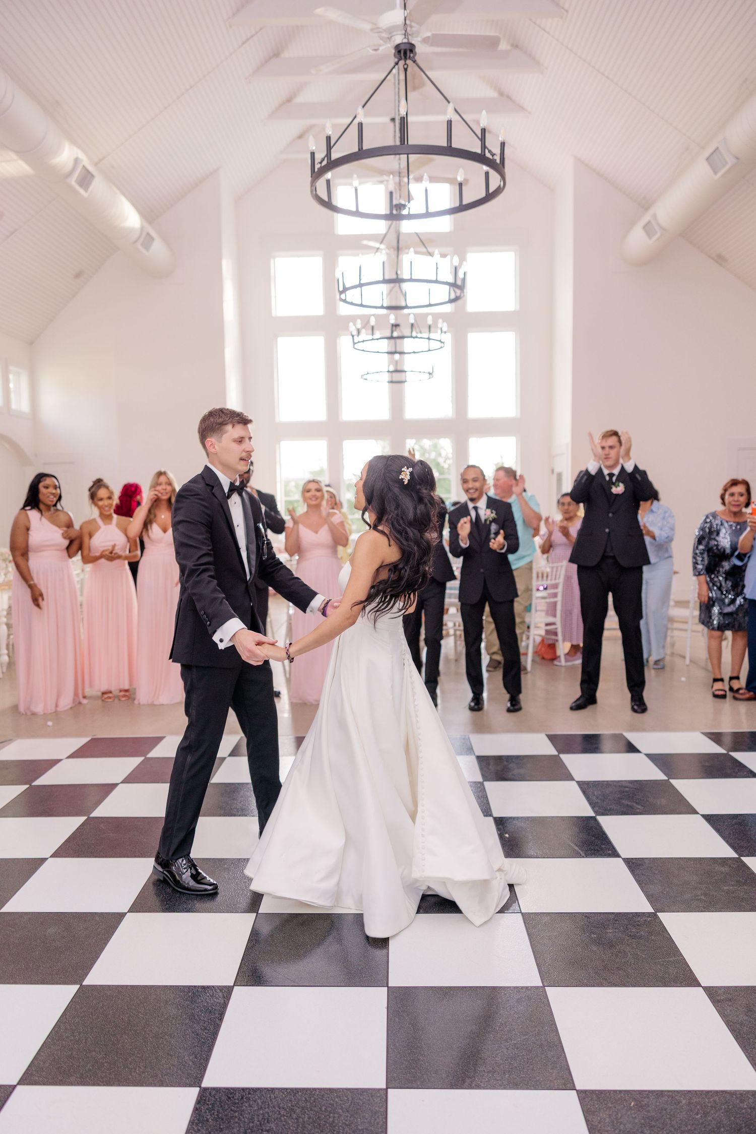First dance on black and white checkered floor under elegant chandeliers.