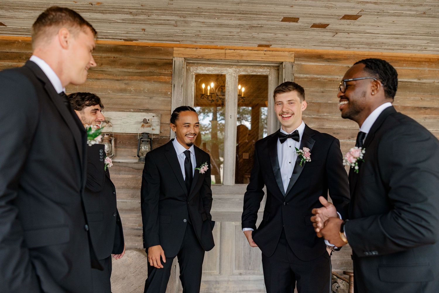 Groomsmen in black tuxedos gathering before wedding ceremony in rustic wood-paneled room.