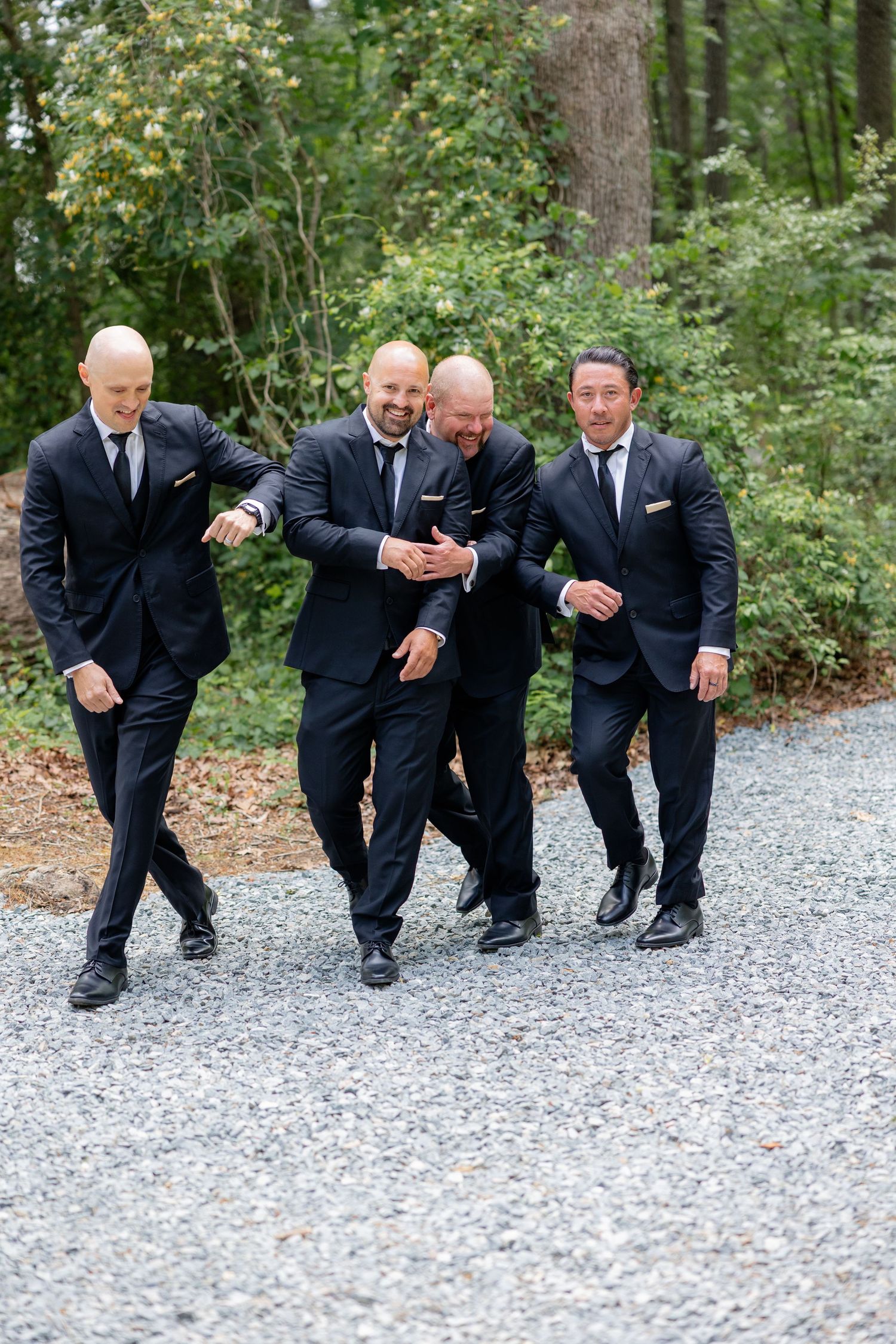 Groomsmen in black suits playfully crouch down on a gravel path during wedding photo session.