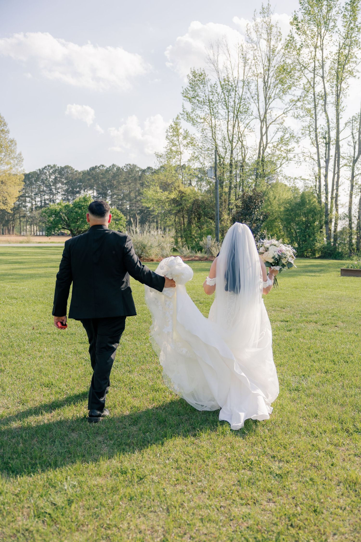 Newlyweds stroll across a sunny lawn with the bride's elegant white dress and veil trailing in the breeze.