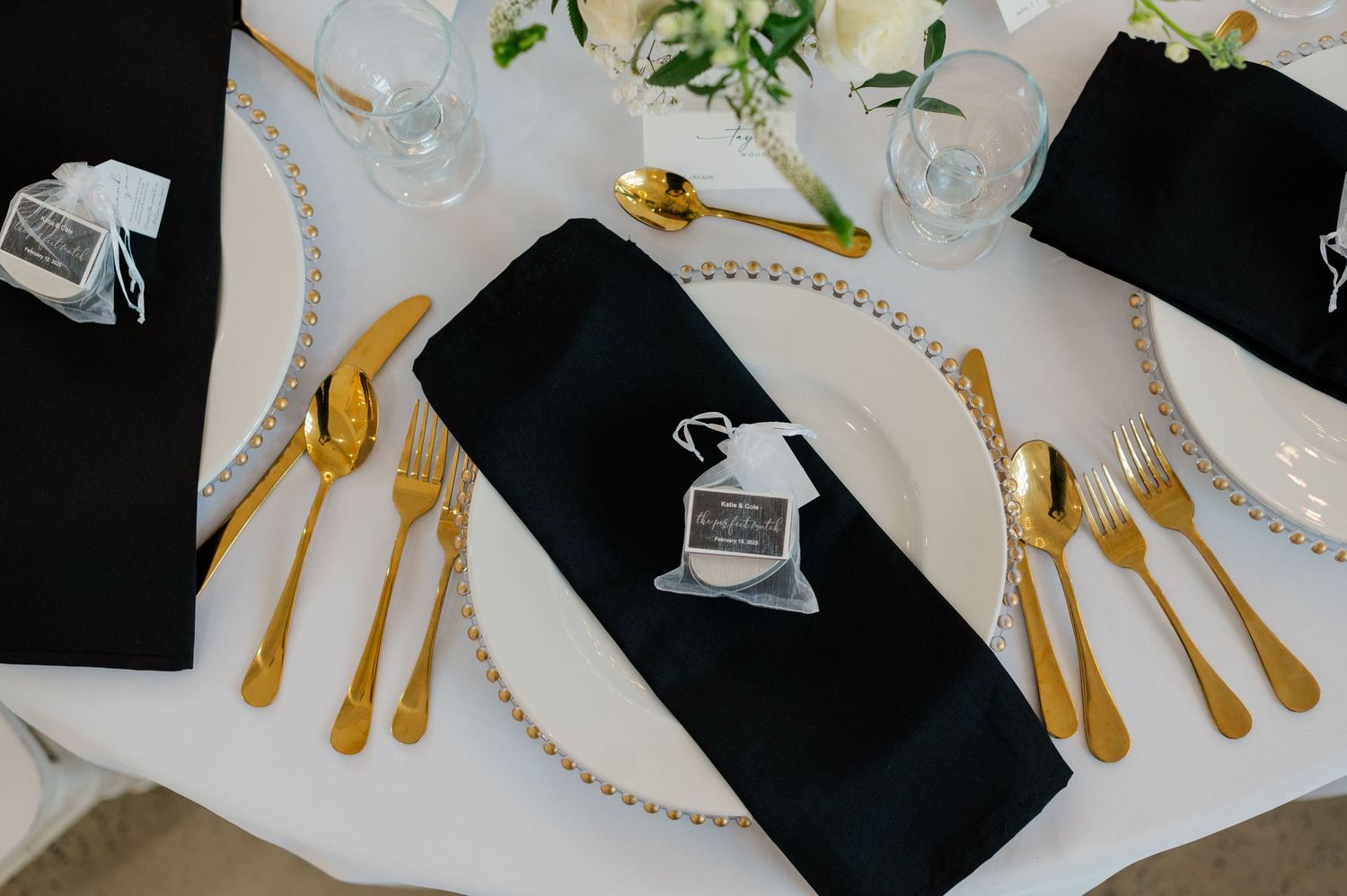 A formal place setting features black napkins, gold cutlery, and small decorative wedding favors on a white tablecloth.