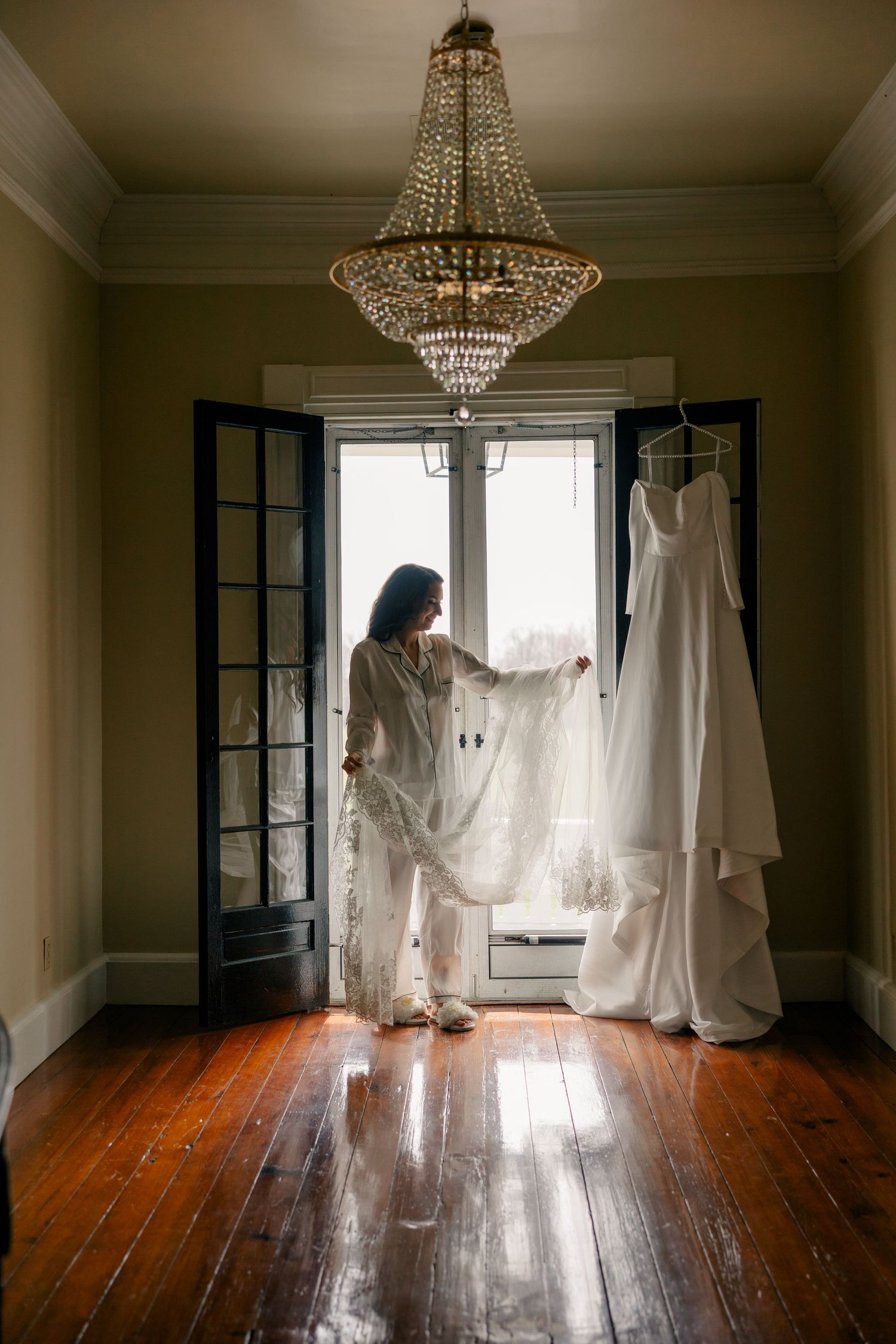 Wedding dress being prepared in a bright room with wood floors and crystal lighting fixtures.