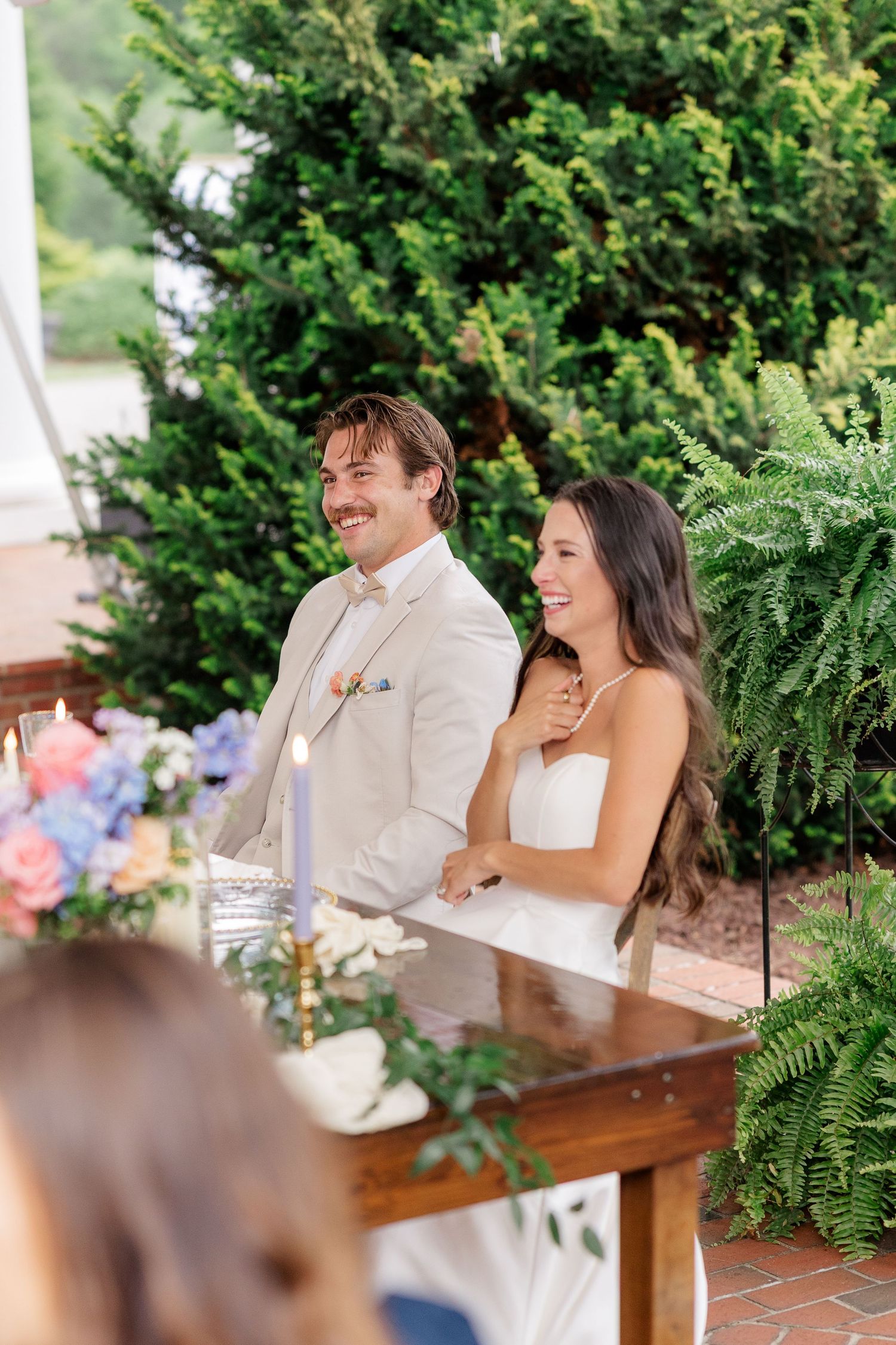A couple shares a joyful moment during their wedding reception while seated at a wooden table surrounded by greenery.