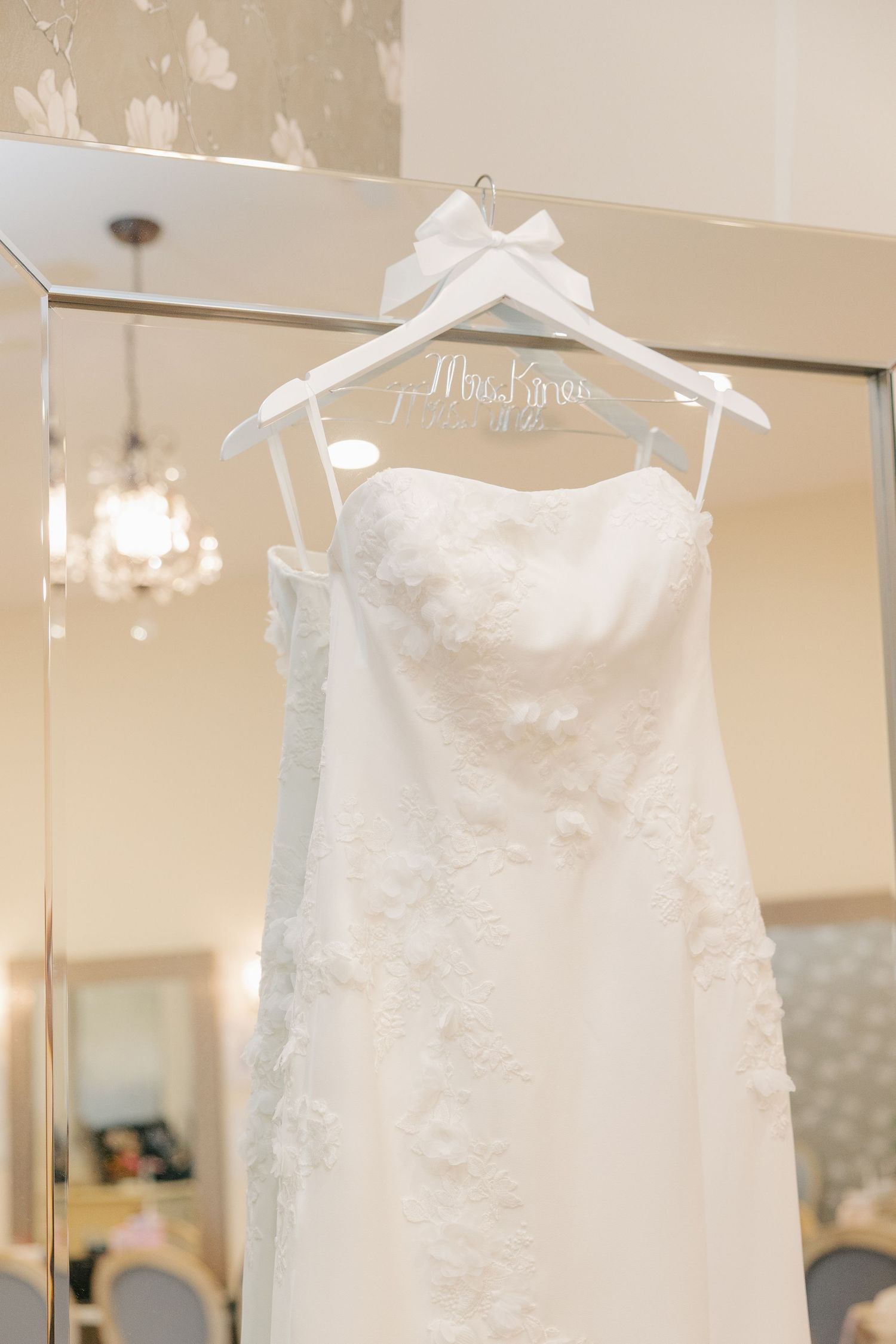 White lace strapless wedding dress hanging on personalized Mrs. hanger in front of elegant mirror.