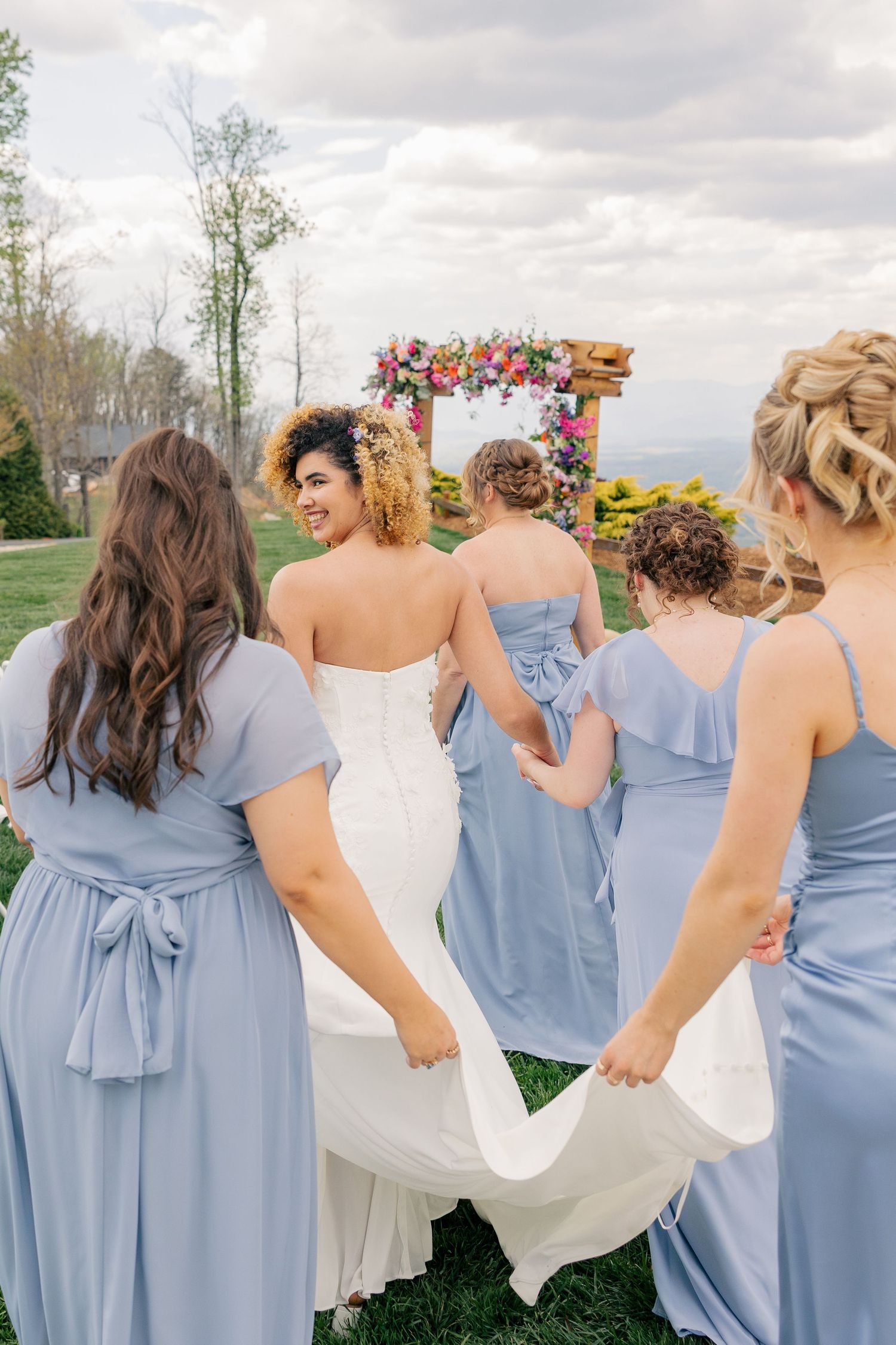 Bride in white gown walks toward floral arch assisted by bridesmaids in dusty blue dresses outdoors.