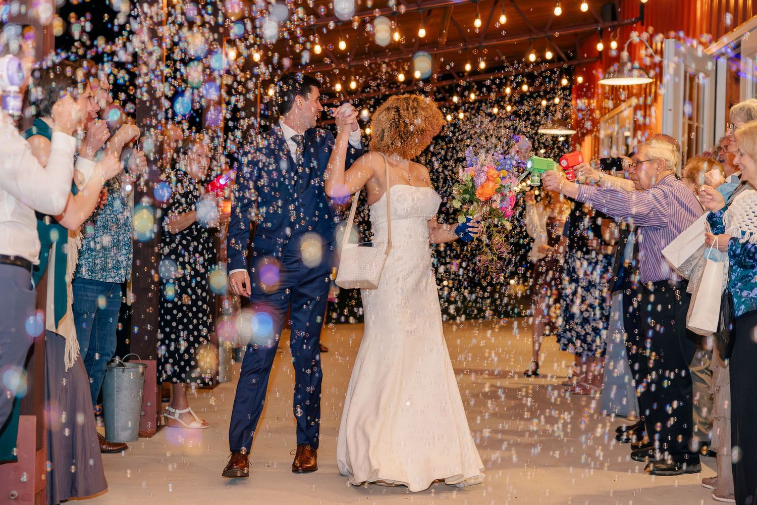 Bride and groom exiting reception through a joyful bubble send-off surrounded by celebrating guests.